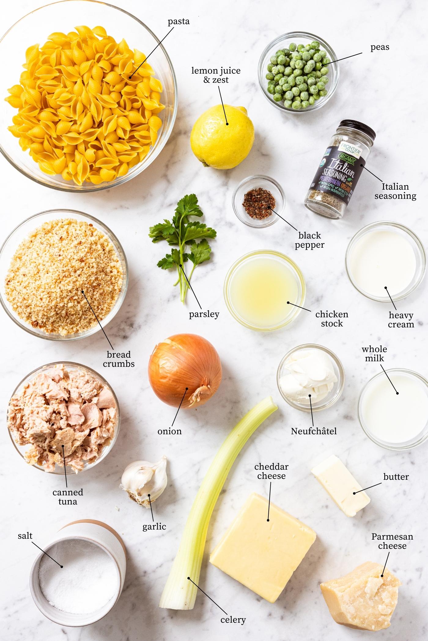 White marble surface filled with all ingredients needed to prepare tuna noodle casserole including pasta, bread crumbs, tuna chunks, celery, onion, milk, cream, spices, and more.
