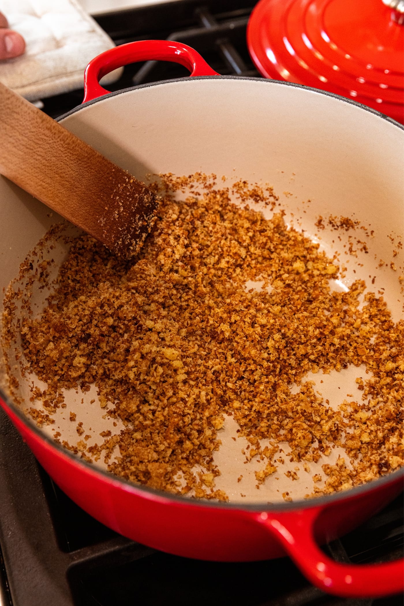Wooden spatula mixing together browned bread crumbs in a large red Dutch oven on the stove.