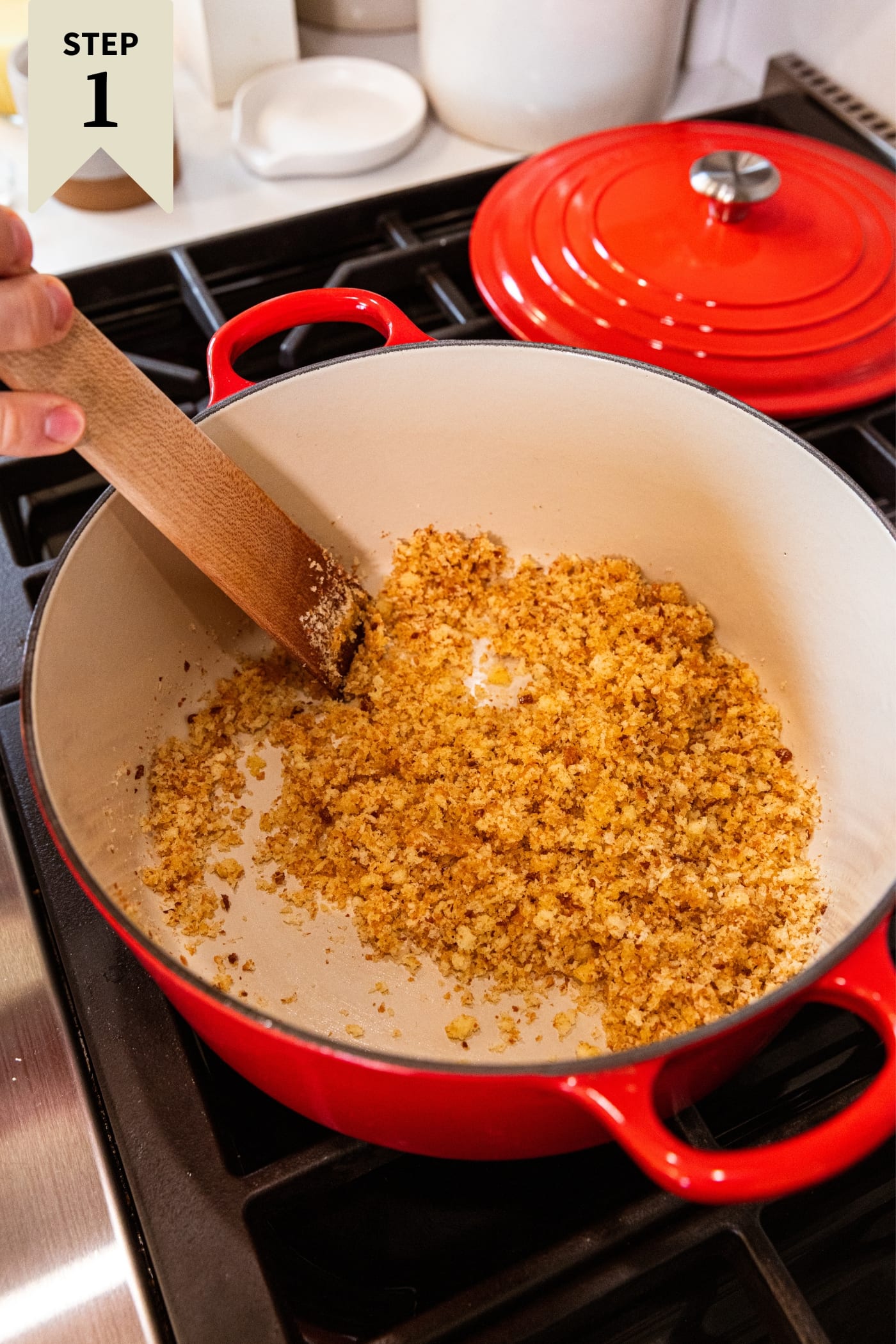 Hand holding wooden spatula mixing bread crumbs in the bottom of a red Dutch oven on the stovetop.