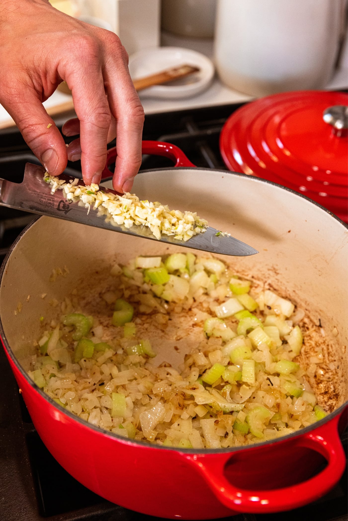 Hand holding knife about to push minced garlic into large red Dutch oven filled with browned celery and onion chopped up.
