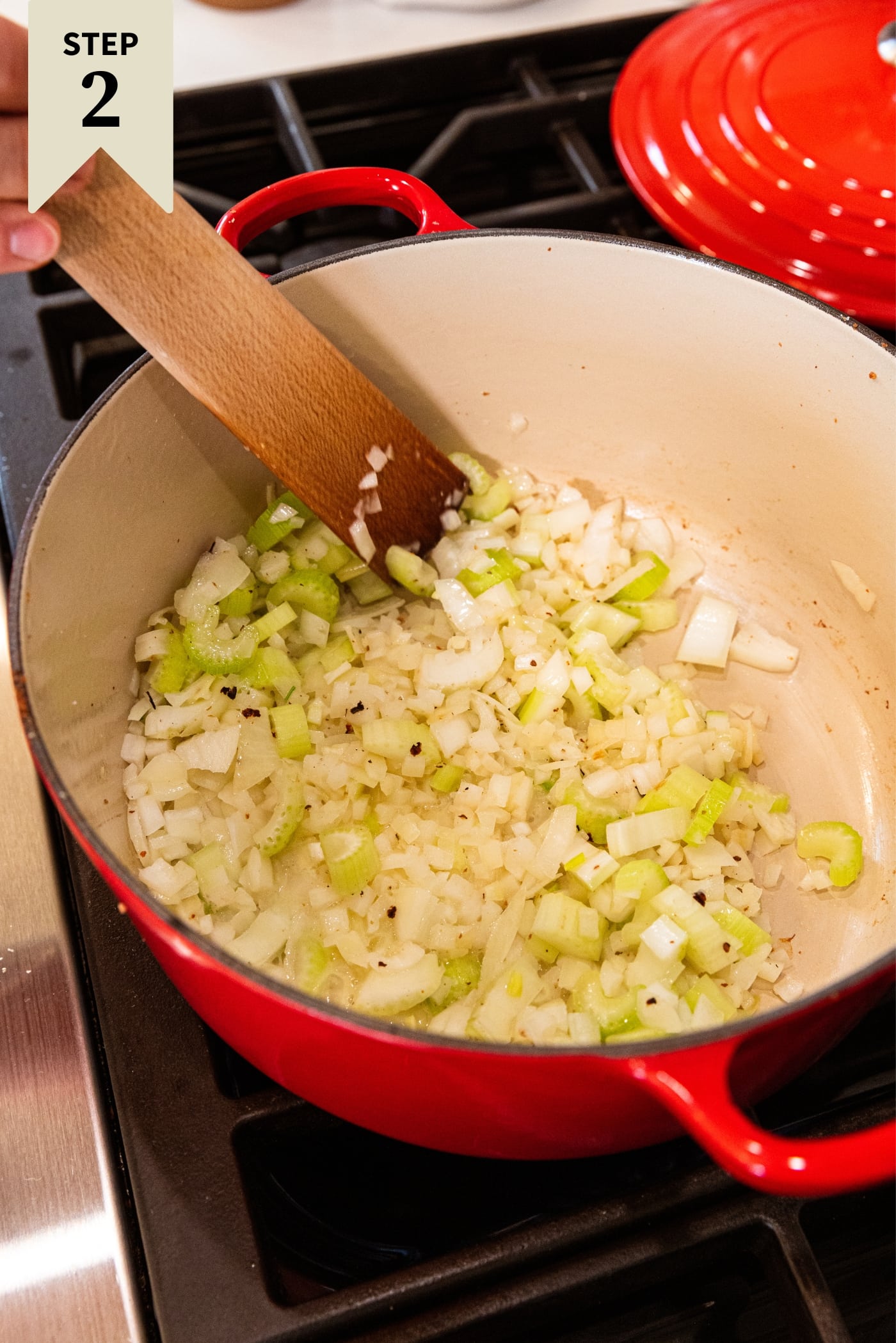 Large red Dutch oven on stovetop with browning chopped celery and onion in bottom with wooden spatula mixing everything together.