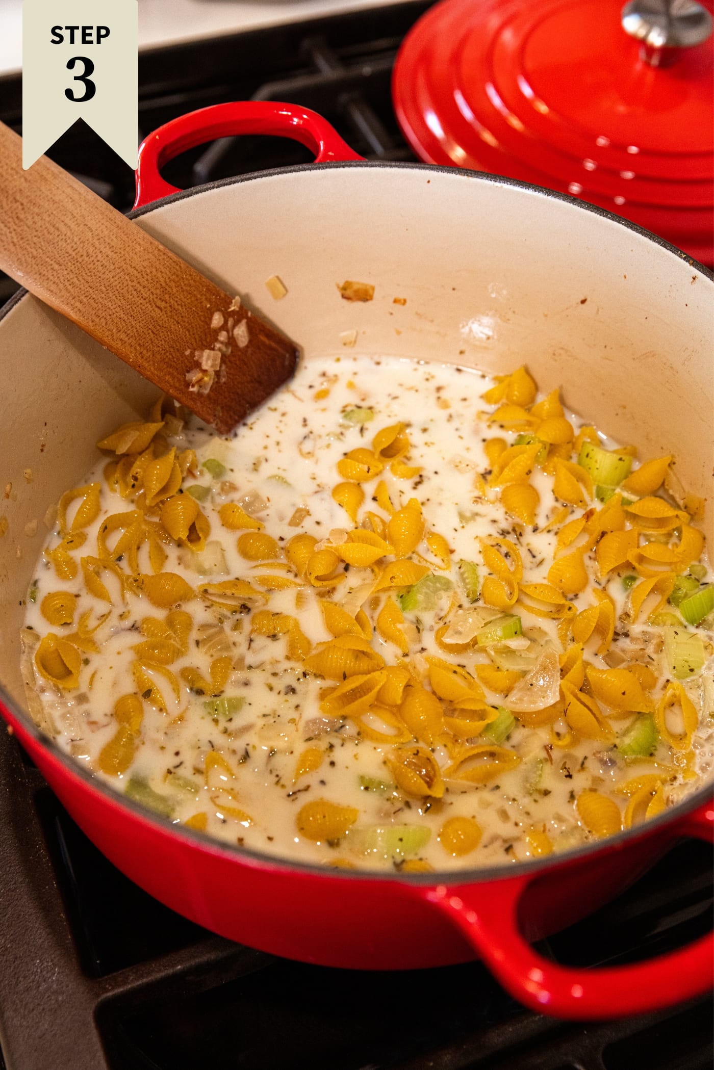 Wooden spatula stirring together uncooked pasta in milk and chicken stock mixture with pieces of chopped and browned onion and celery showing through.
