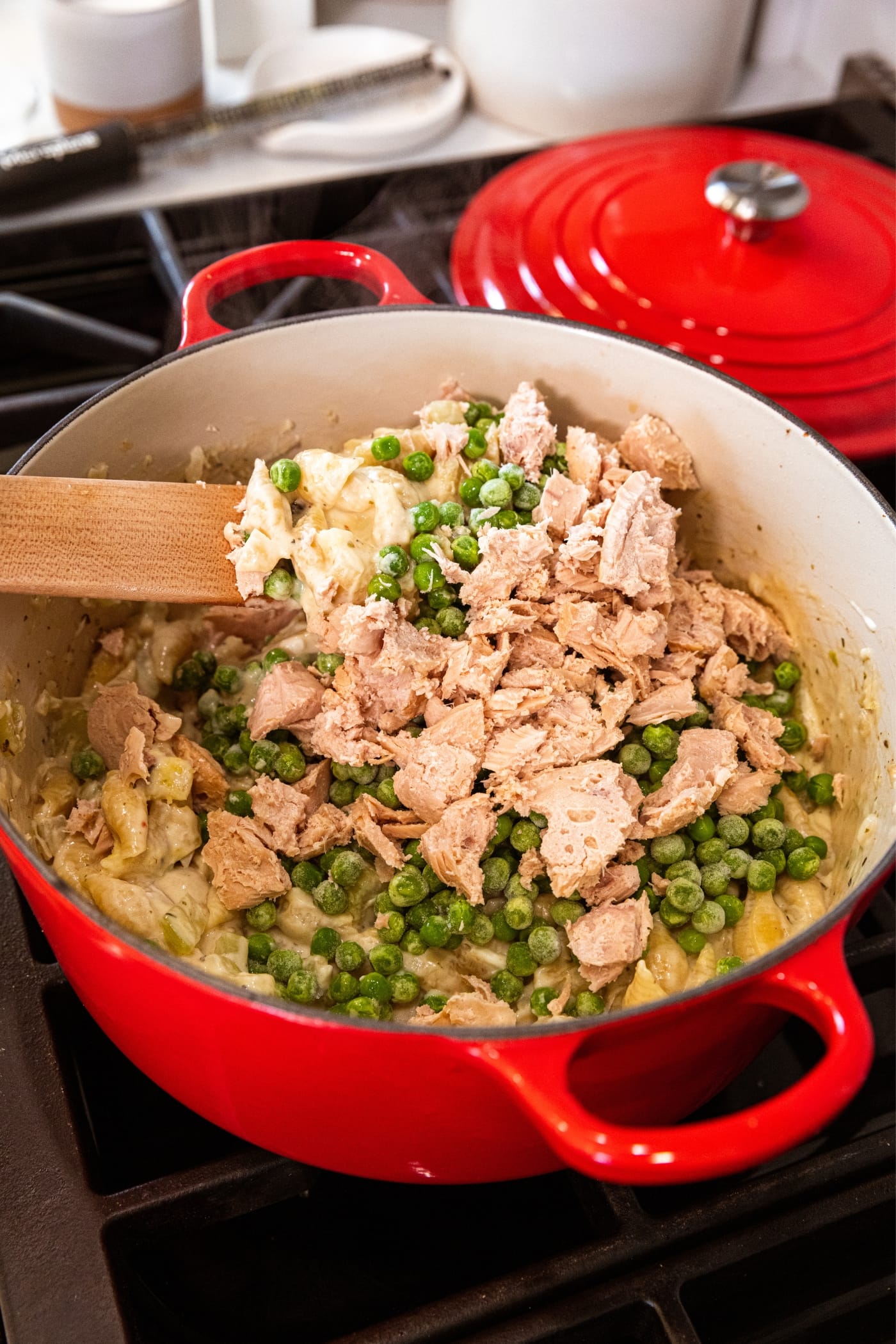 Wooden spatula mixing together chunks of tuna with peas and pasta in large red Dutch oven.