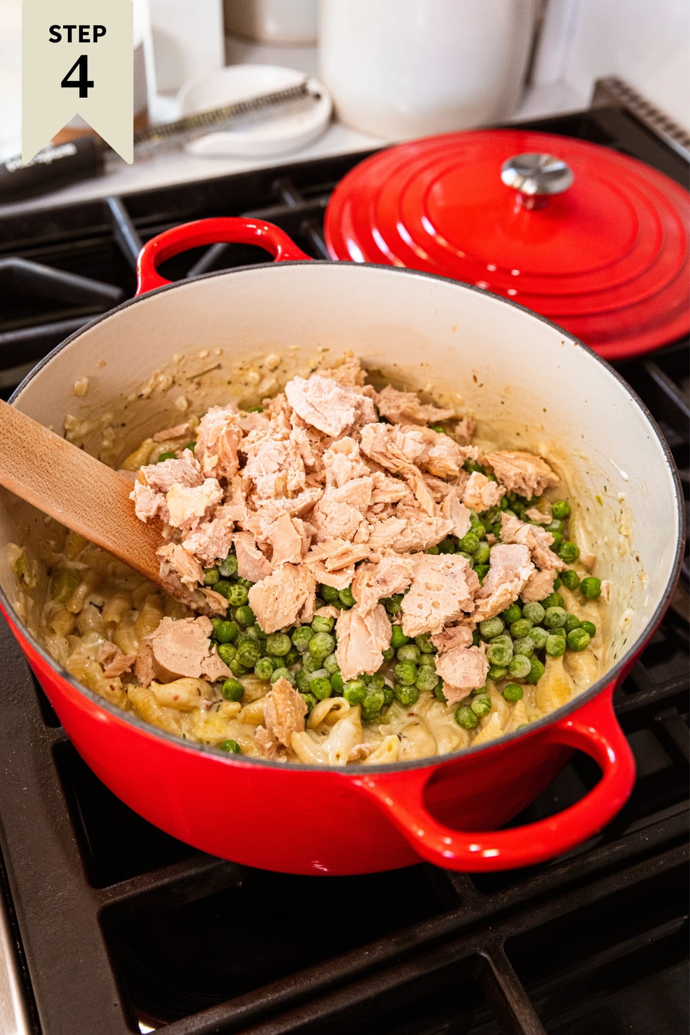 Chunks of tuna and peas sitting on top of cooking pasta in large red Dutch oven on stovetop.