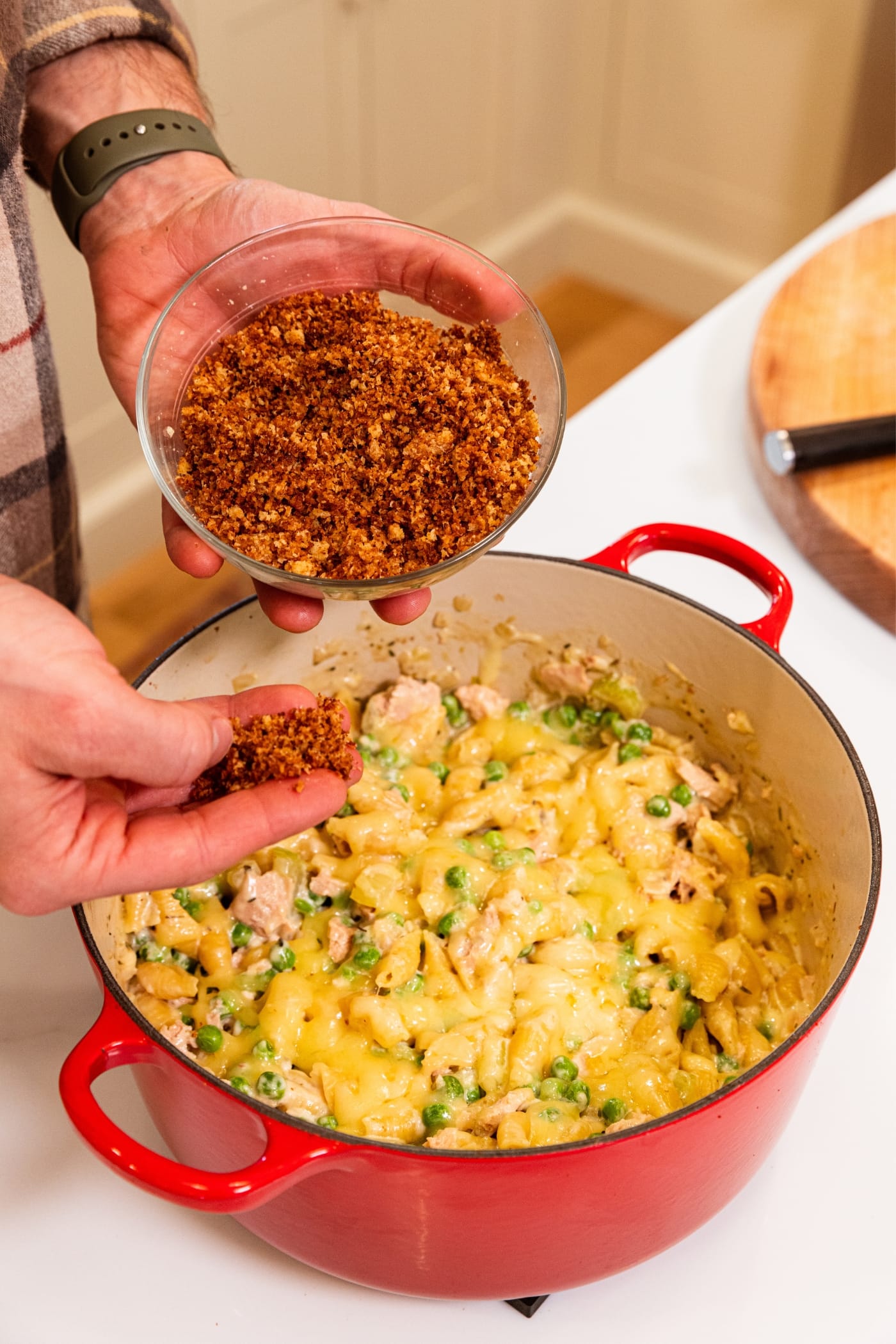 Hand holding small glass bowl filled with browned bread crumbs about to sprinkle over the top of tuna noodle casserole in large red Dutch oven.