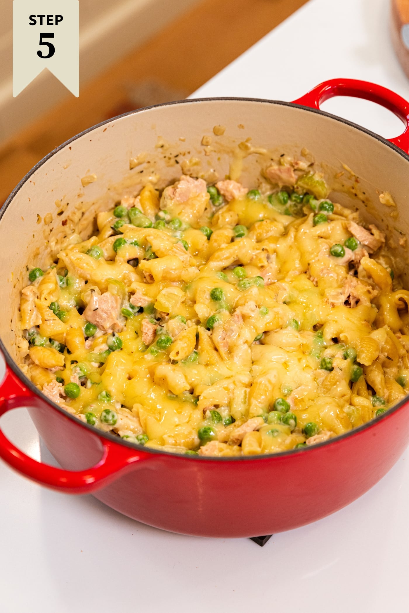 Large red Dutch oven sitting on white surface with tuna noodle casserole with melted cheese inside with peas and chunks of tuna showing through.