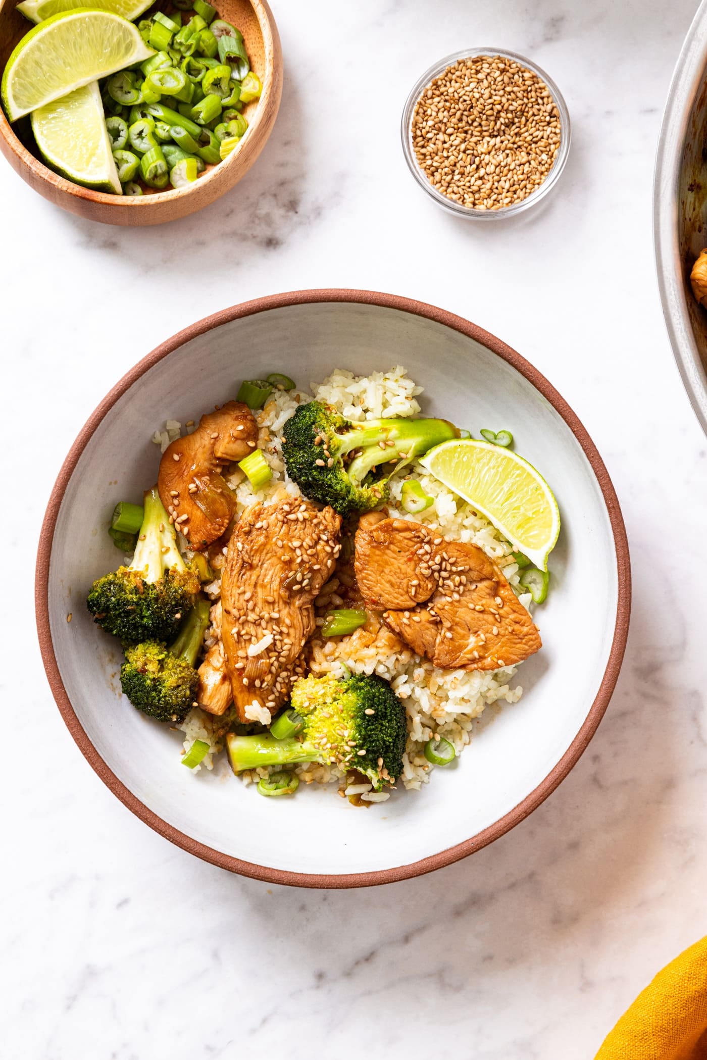 White marble surface with bowl filled with rice and sesame chicken and broccoli on top with small bowl filled with toasted sesame seeds and another bowl filled with scallions and lime wedges.