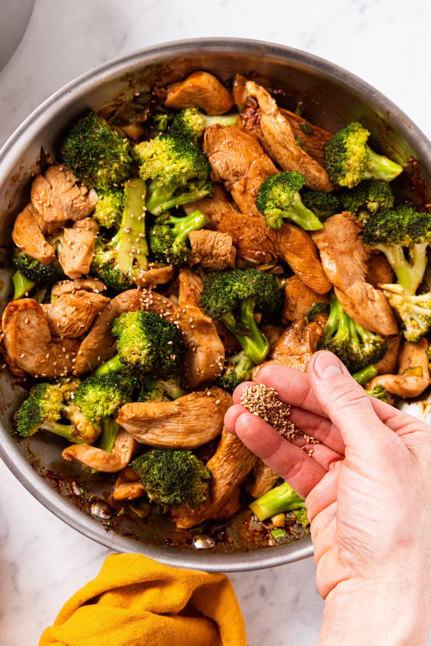 Hand holding sesame seeds about to sprinkle them on top of a large skillet filled with cooked sesame chicken and broccoli tossed in a soy sauce.
