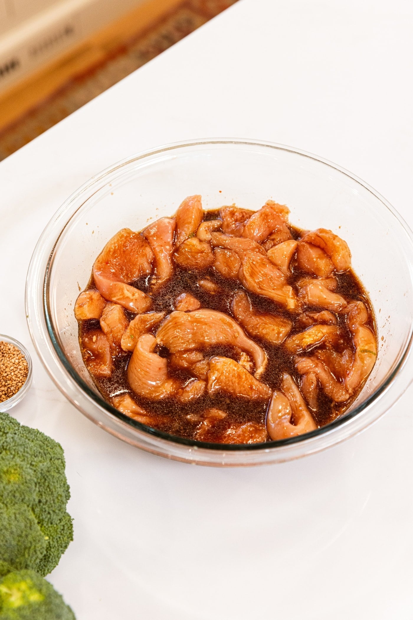 Glass bowl filled with raw chicken in brown soy sauce marinade sitting on white countertop before being put in the refrigerator.