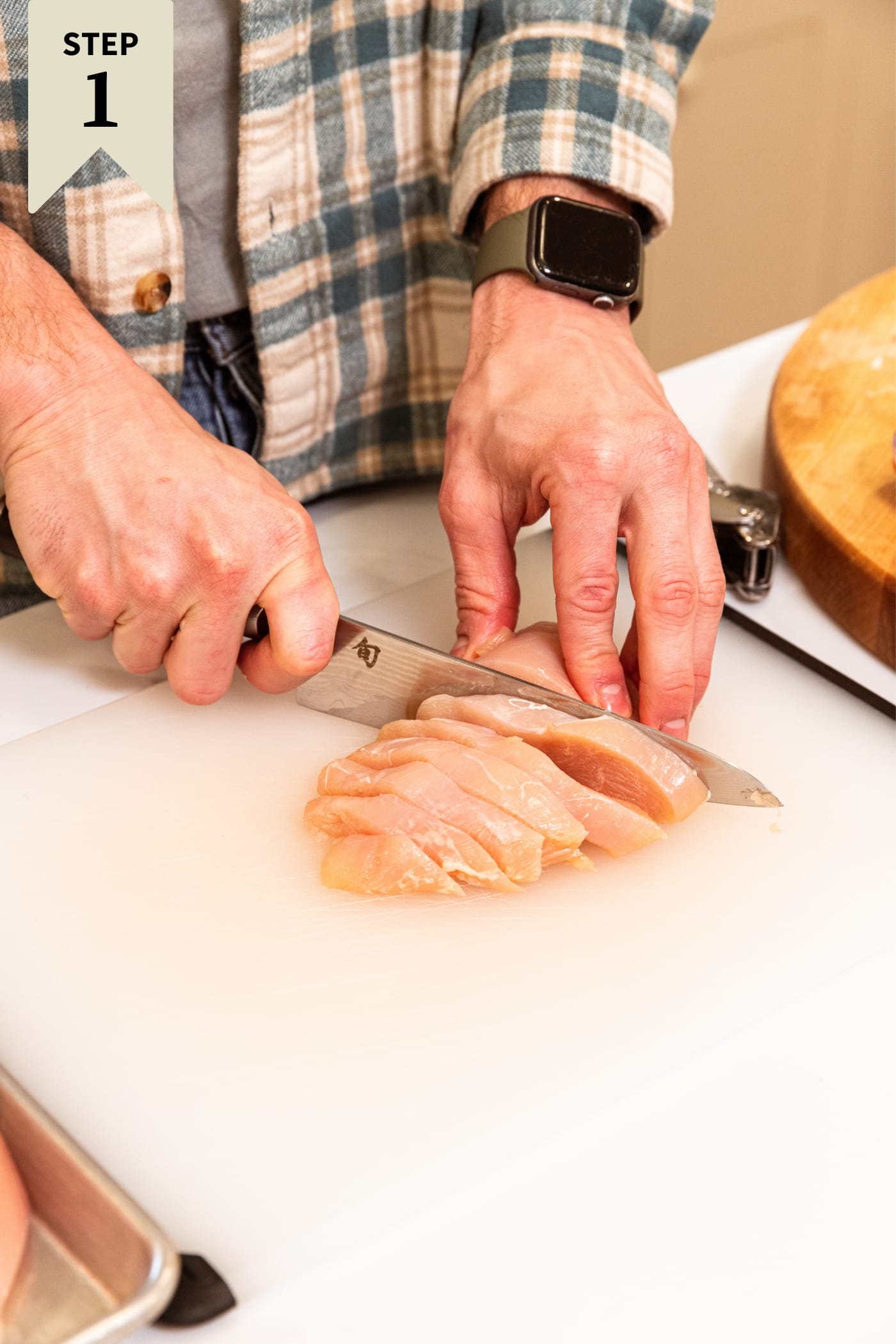Hand using knife to slice up chicken breast into slices on plastic cutting board with wood cutting board in background.