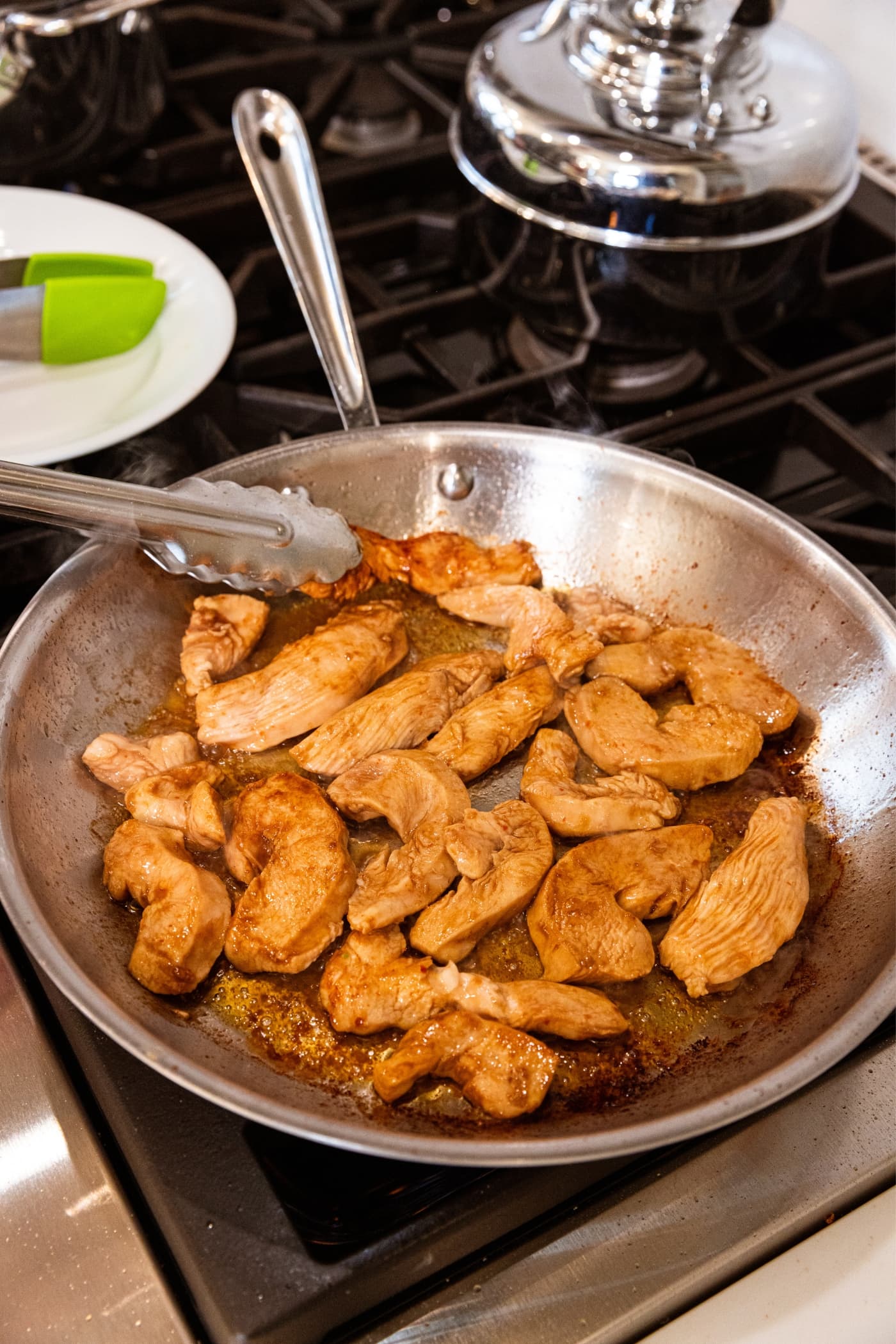 Cooked chicken in brown sauce in metal skillet on stovetop with tongs removing pieces onto a white plate to the side.