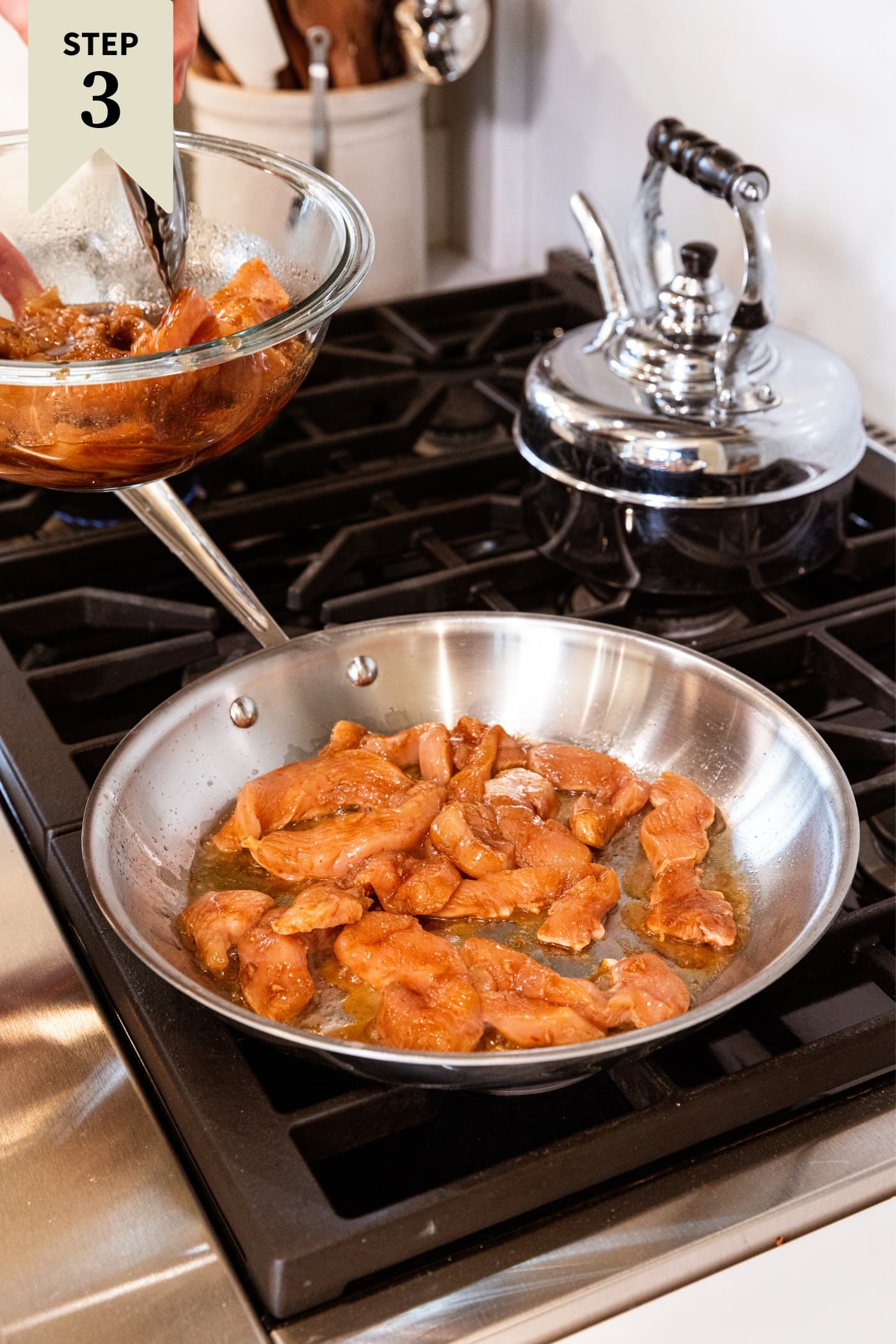 Metal skillet sitting on stovetop filled with cooking chicken with hand using tongs to put more chicken into skillet to cook.