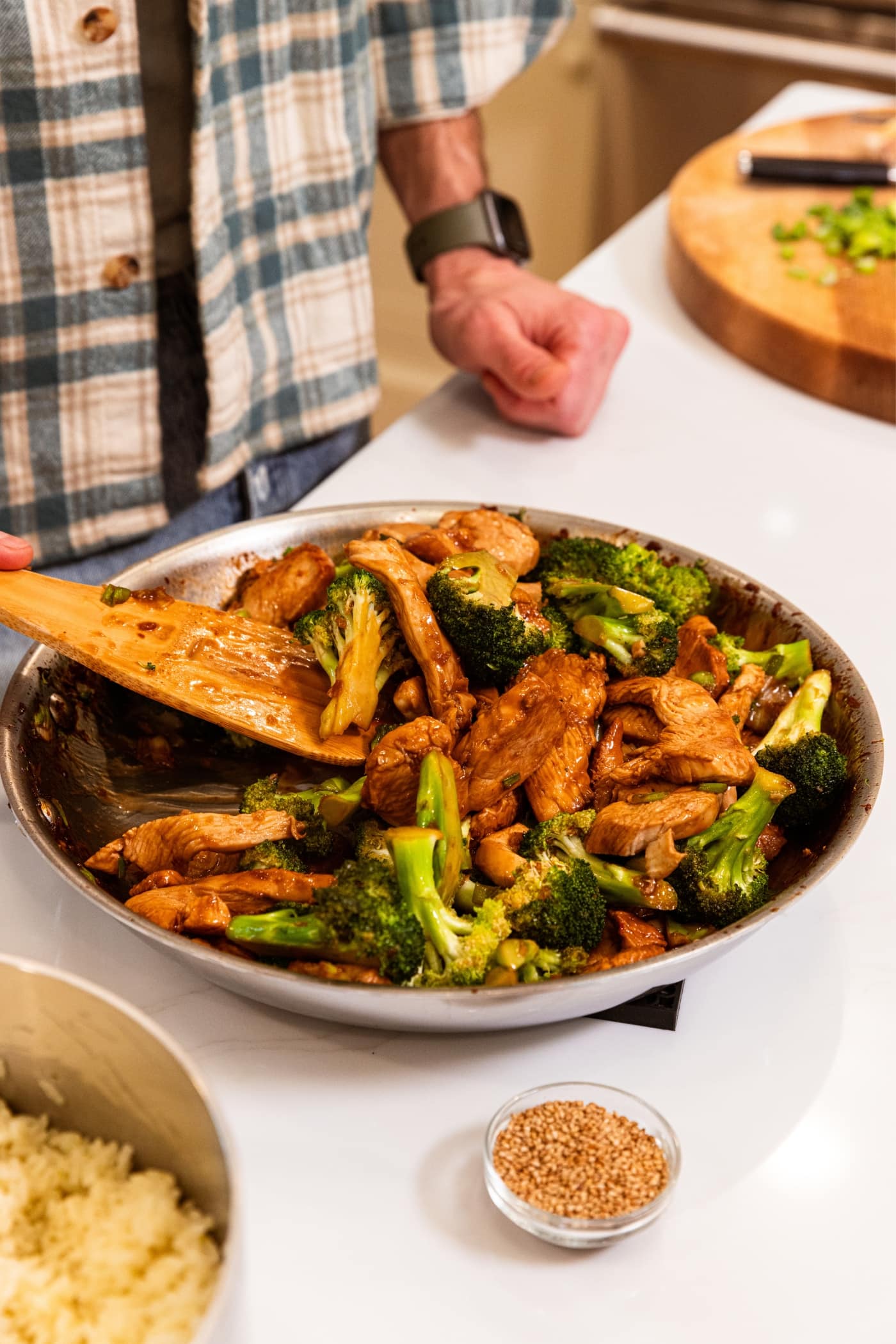 Hand using wooden spatula to mix together pieces of cooked chicken with broccoli florets in soy sauce all in metal skillet on white surface.
