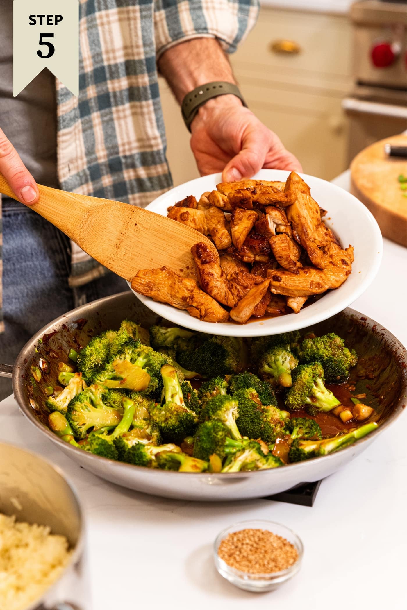Hand holding wooden spatula about to push cooked pieces of chicken into a skillet filled with broccoli florets tossed in soy sauce all on white surface.
