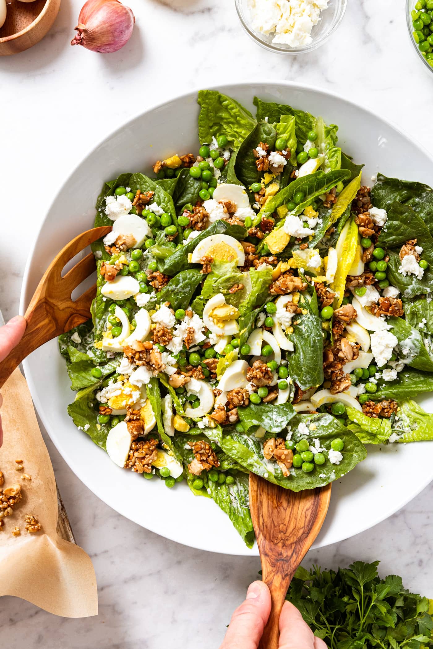 Two hands holding wooden tongs scooping up serving of salad from large white serving platter filled with spring pea salad topped with peas, feta cheese, hardboiled eggs, and creamy dressing, all on white marble surface.