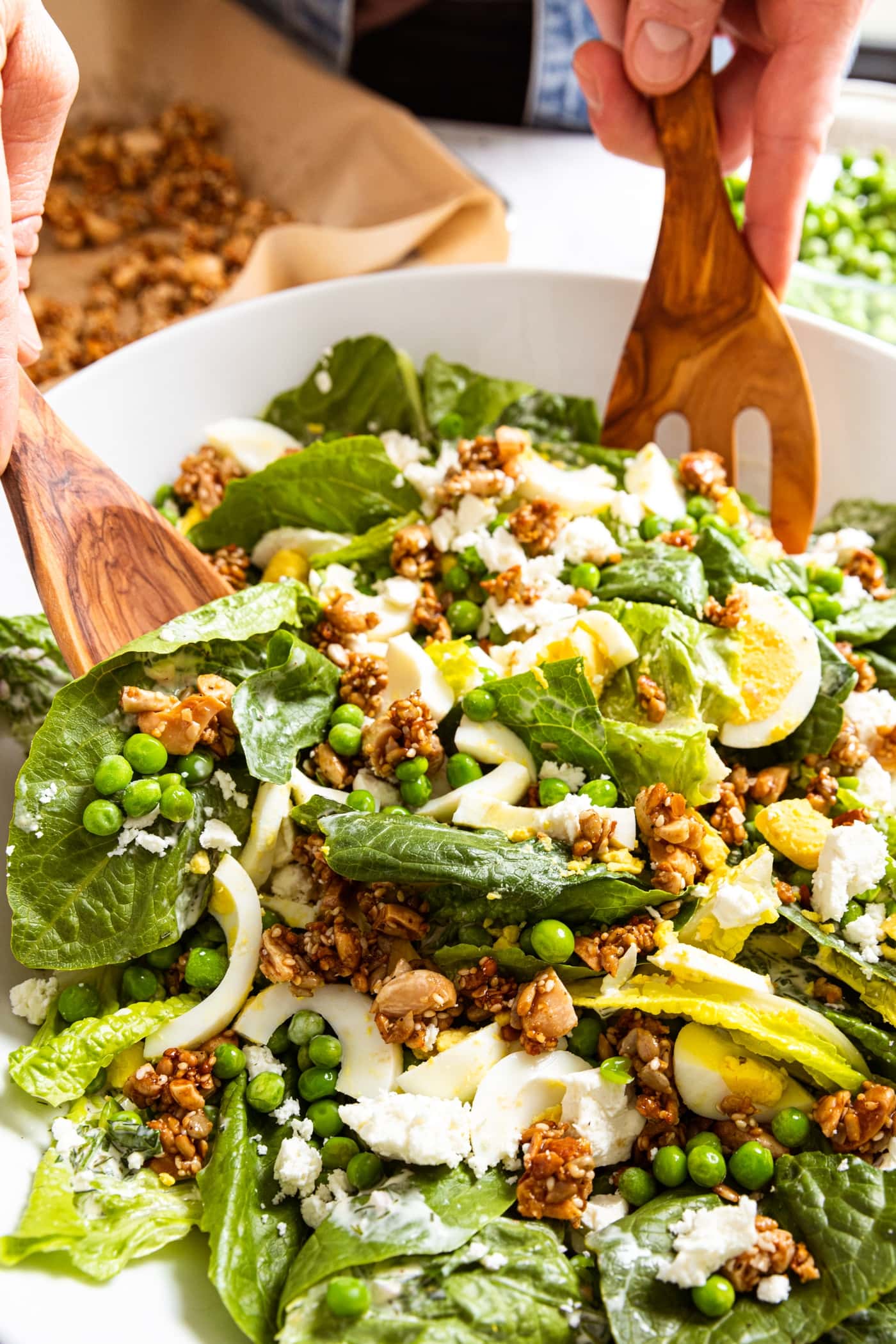 Two wooden tongs pulling up a serving a spring pea salad from large white serving platter topped with crunchy nut topping and hardboiled eggs and feta cheese.