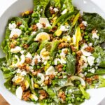 Large white serving platter sitting on white marble surface with lettuce, hardboiled eggs, nut topping, and creamy dressing all over the salad.
