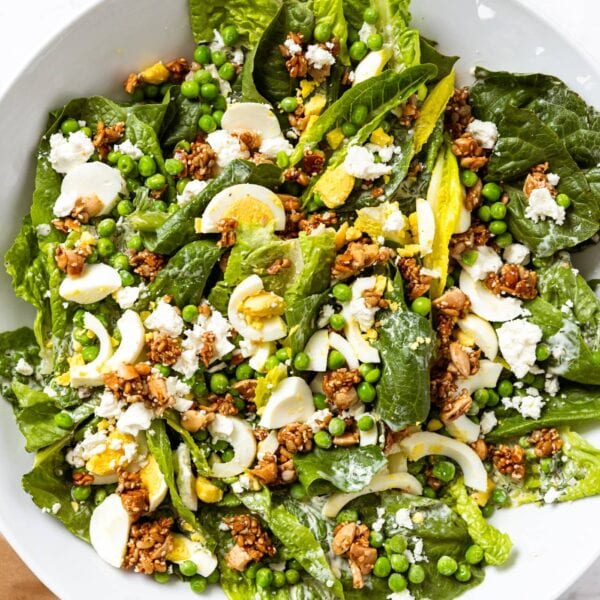 Large white serving platter sitting on white marble surface with lettuce, hardboiled eggs, nut topping, and creamy dressing all over the salad.