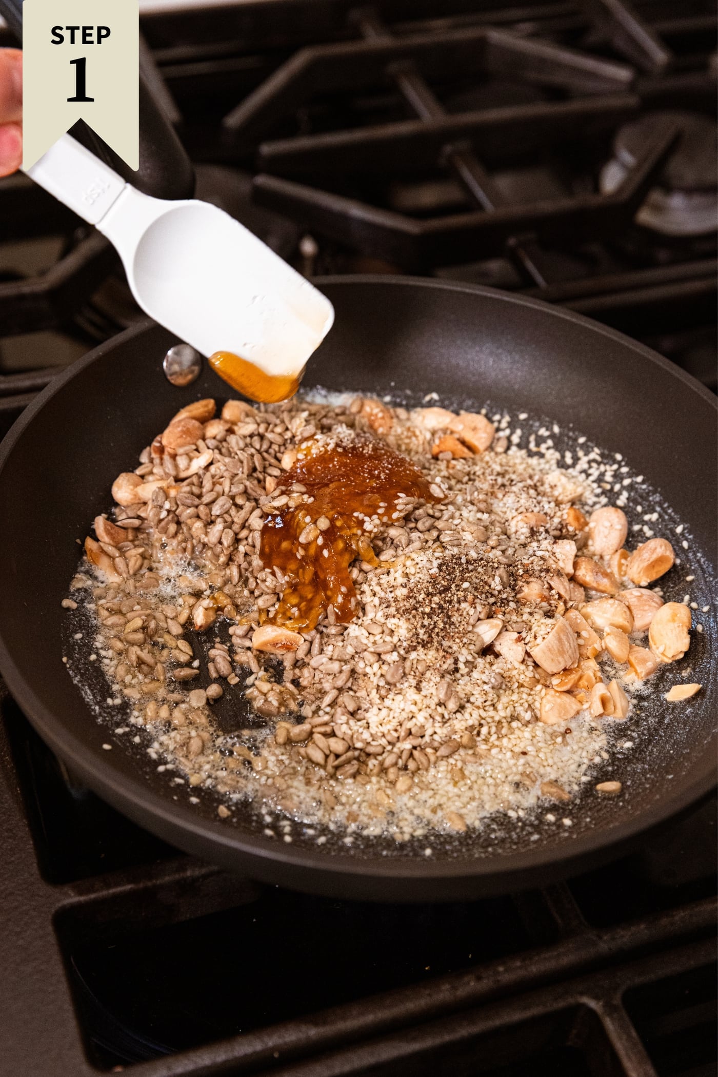 Black skillet filled with sesame seeds, sunflower seeds, honey, and chopped Marcona almonds right before a white spatula mixes everything together over the heat.