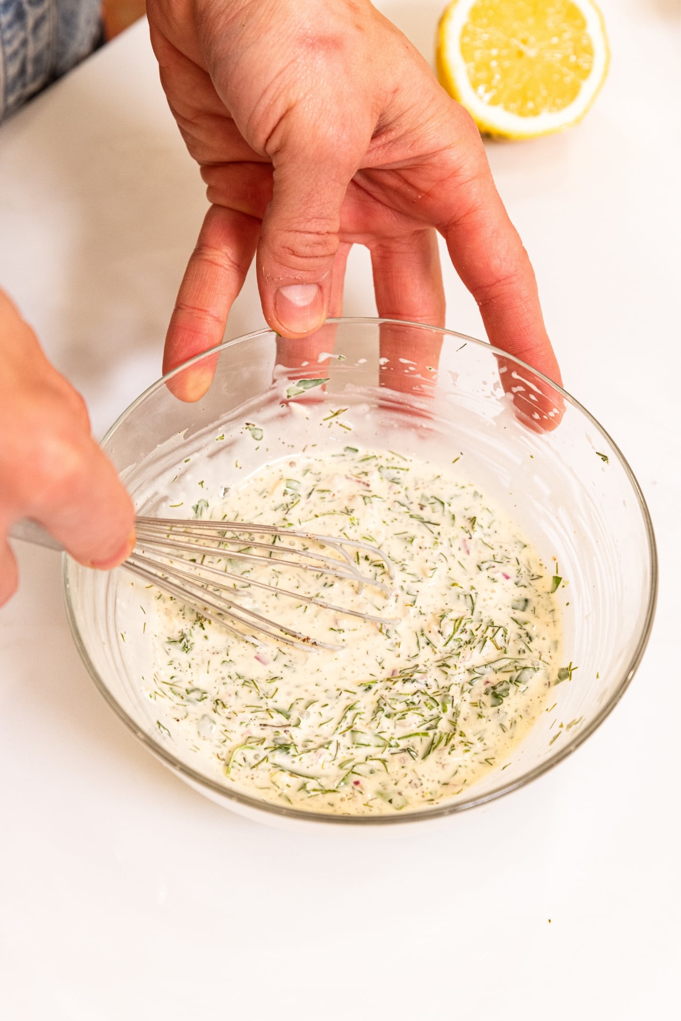 Hands holding onto the sides of a glass bowl filled with white creamy salad dressing with whisk mixing everything together.