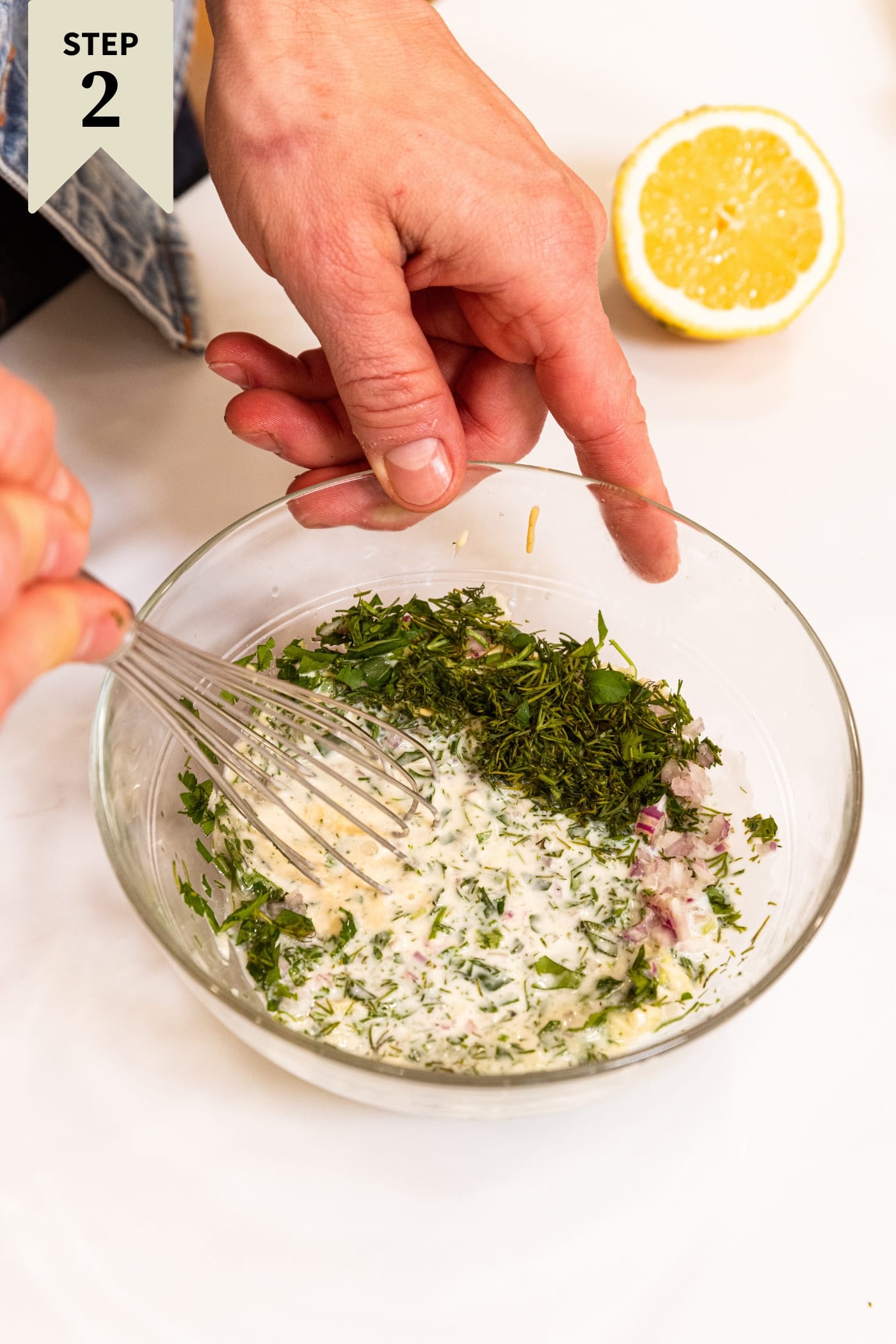 Small glass bowl filled with ingredients needed to prepare salad dressing including buttermilk, herbs, and mustard with whisk about to mix everything together.