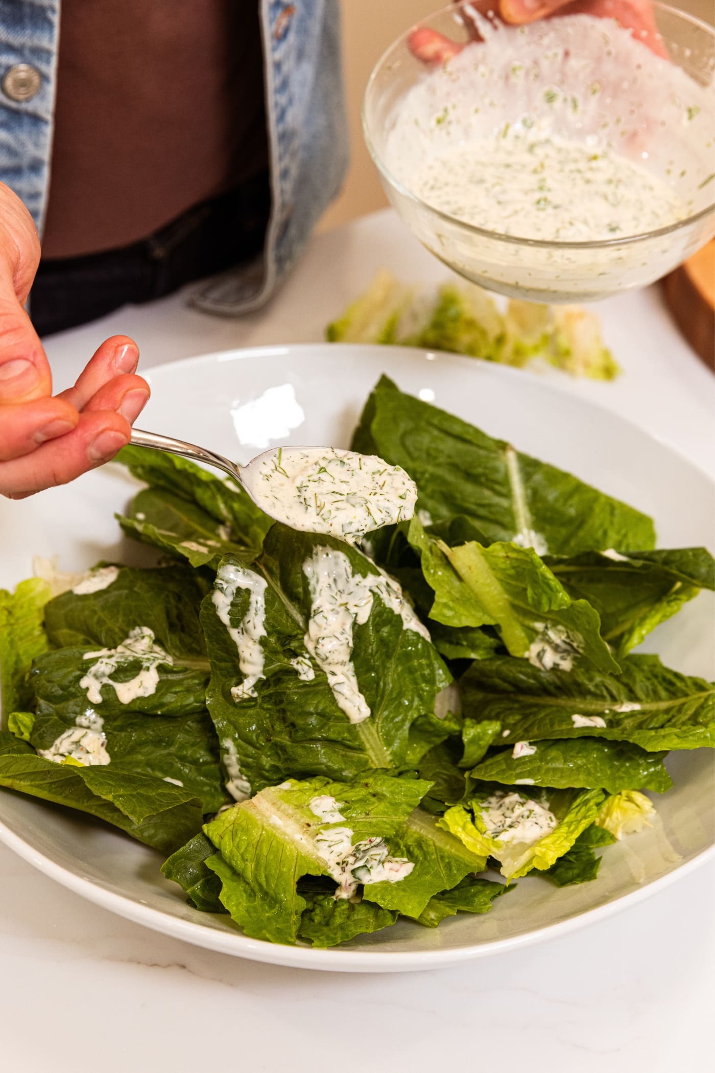 Hand holding spoon filled with creamy white salad dressing drizzling it on top of lettuce in large white platter.