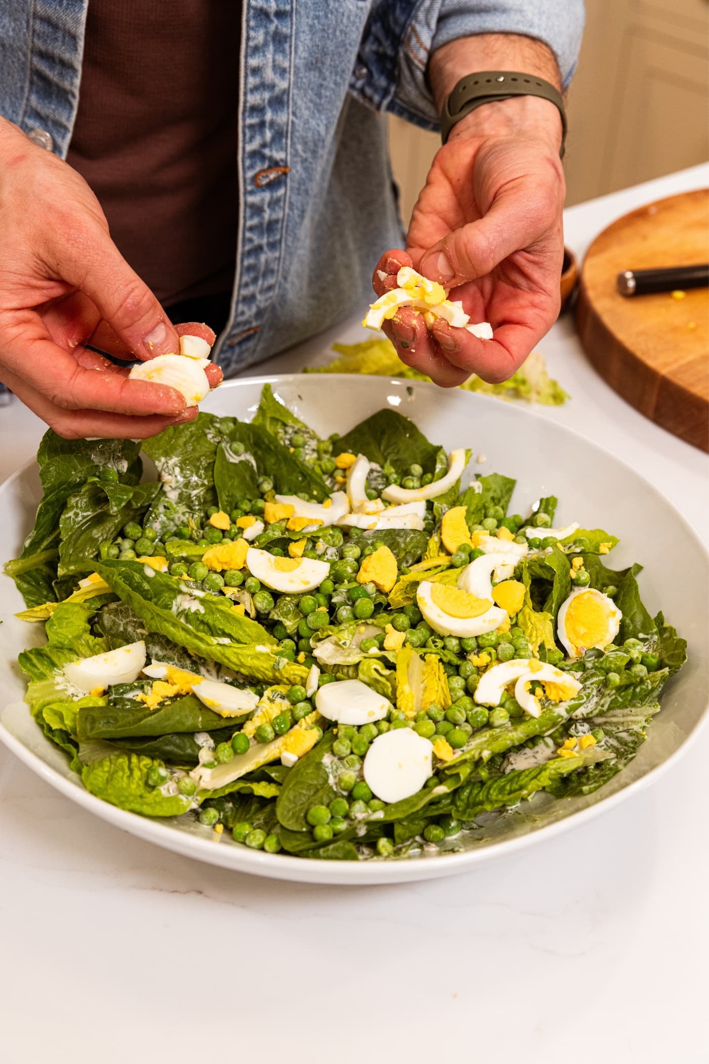 Two hands holding crumbled up slices of hardboiled eggs sprinkling them on top of a salad with lettuce and peas in large white platter.