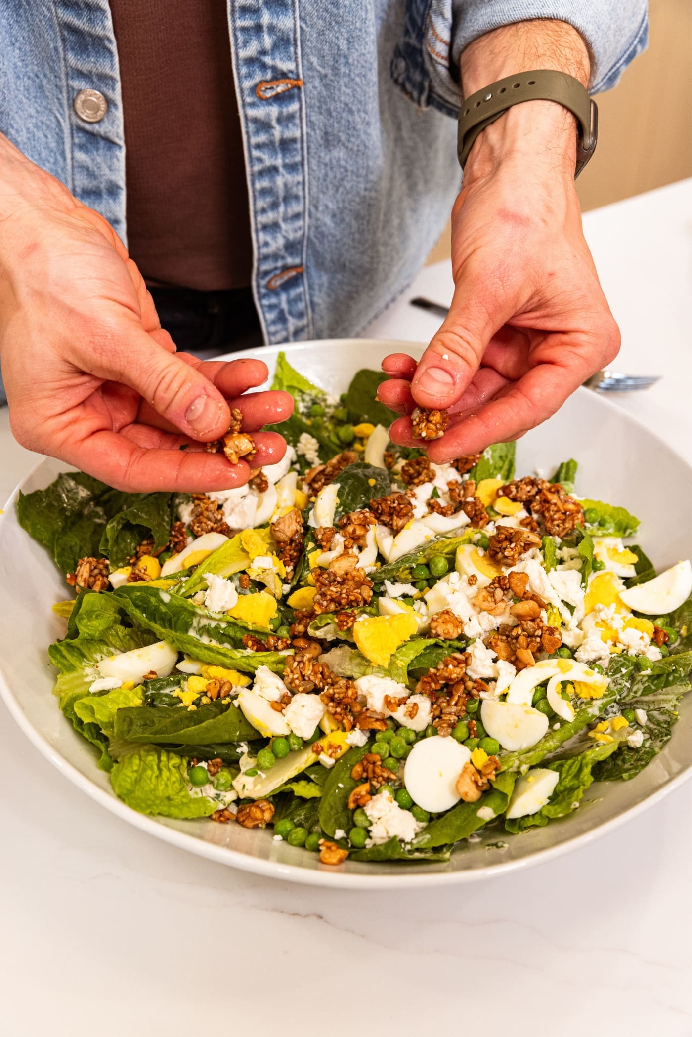Two hands sprinkling crunchy nut and seed topping on top of salad with green lettuce, hardboiled eggs, peas, and feta cheese in large white serving platter.