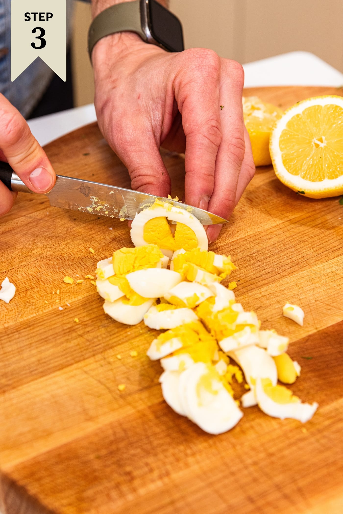 Hand using knife to slice hardboiled eggs into slices on a wood cutting board with interior yellow yolks showing.