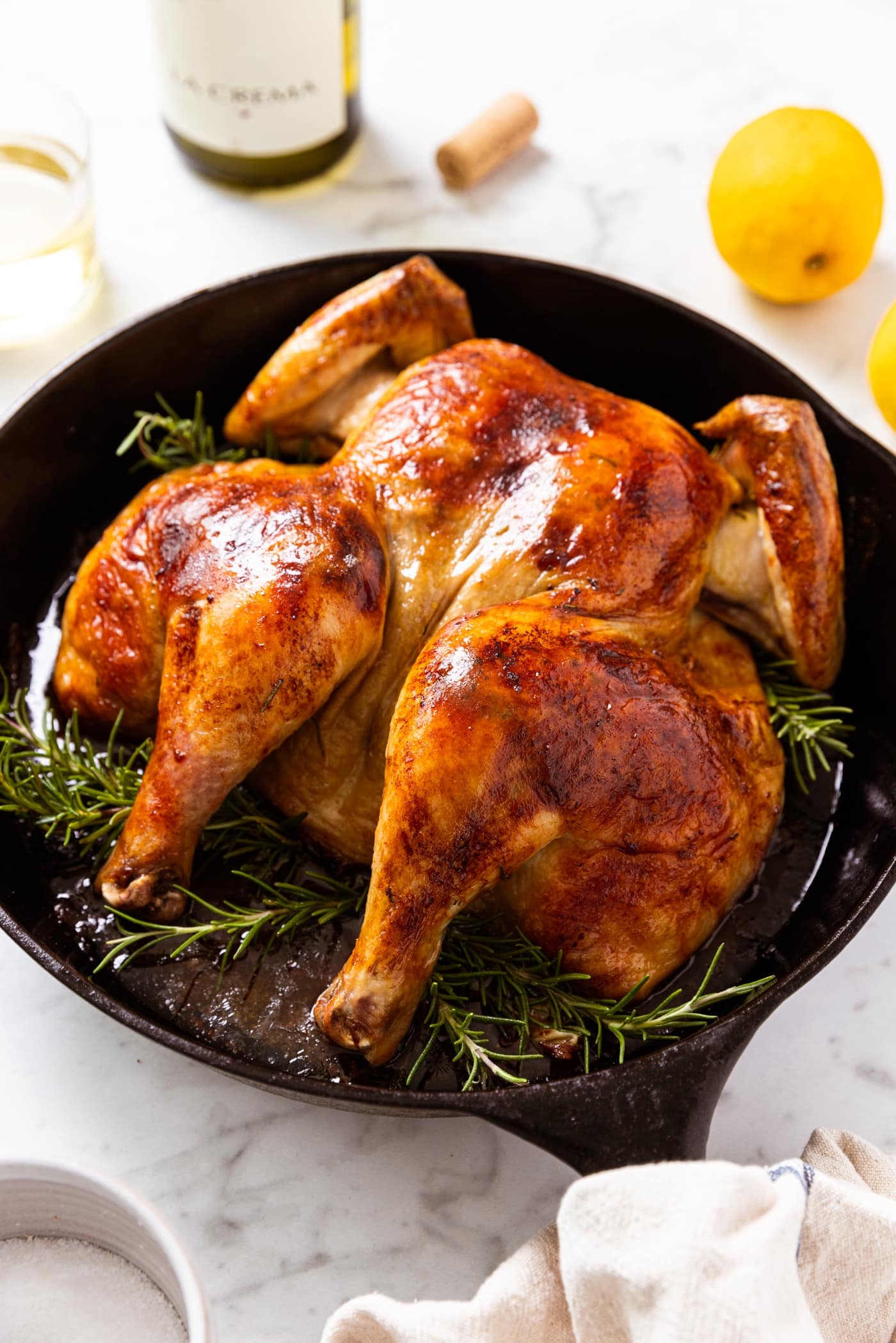 Black skillet filled with roasted chicken with golden top with herbs all around the base of the chicken all on white marble surface.
