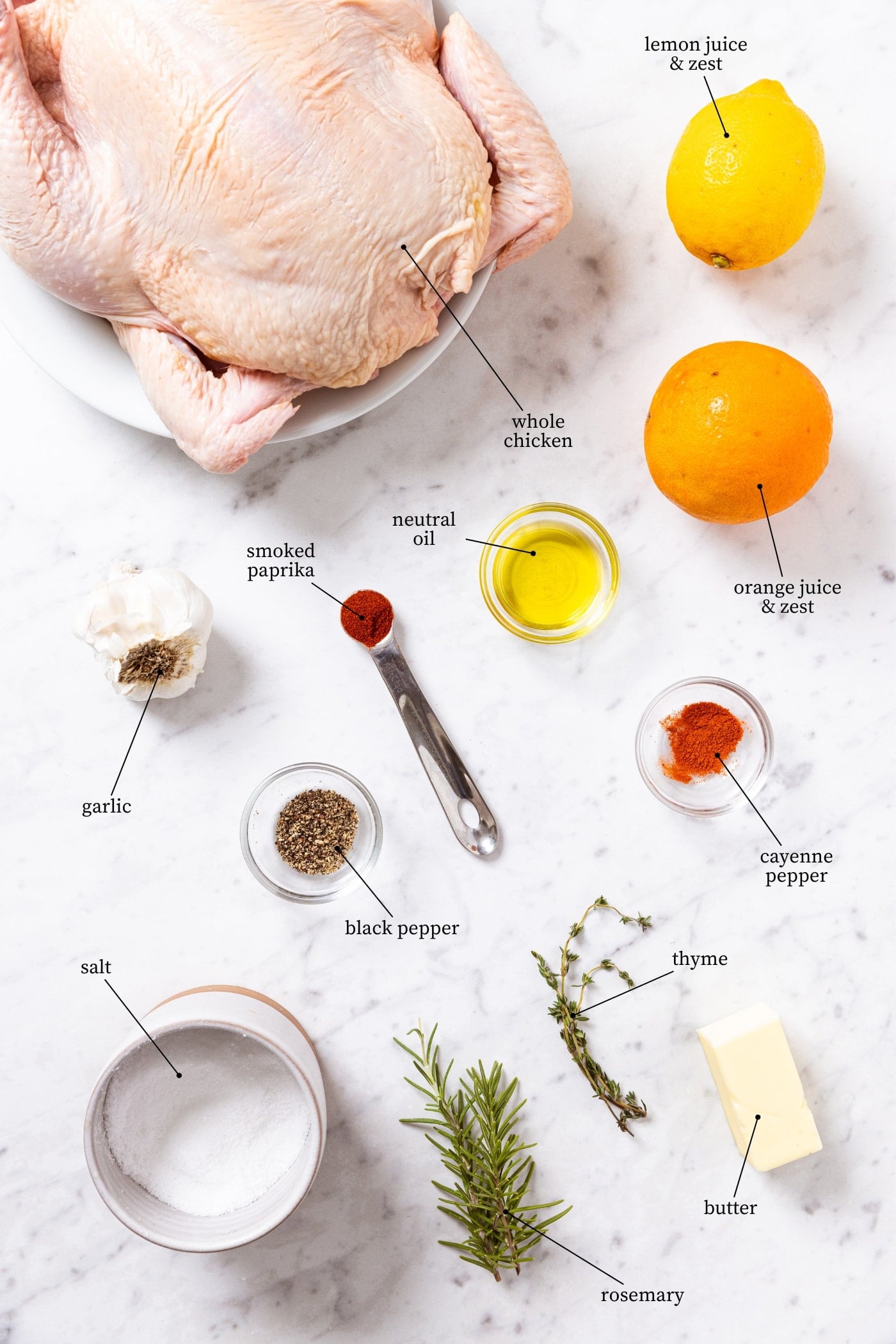 White marble surface filled with all ingredients needed to make citrus and herb roast chicken including raw whole chicken, spices, salt, orange, lemon, and more.