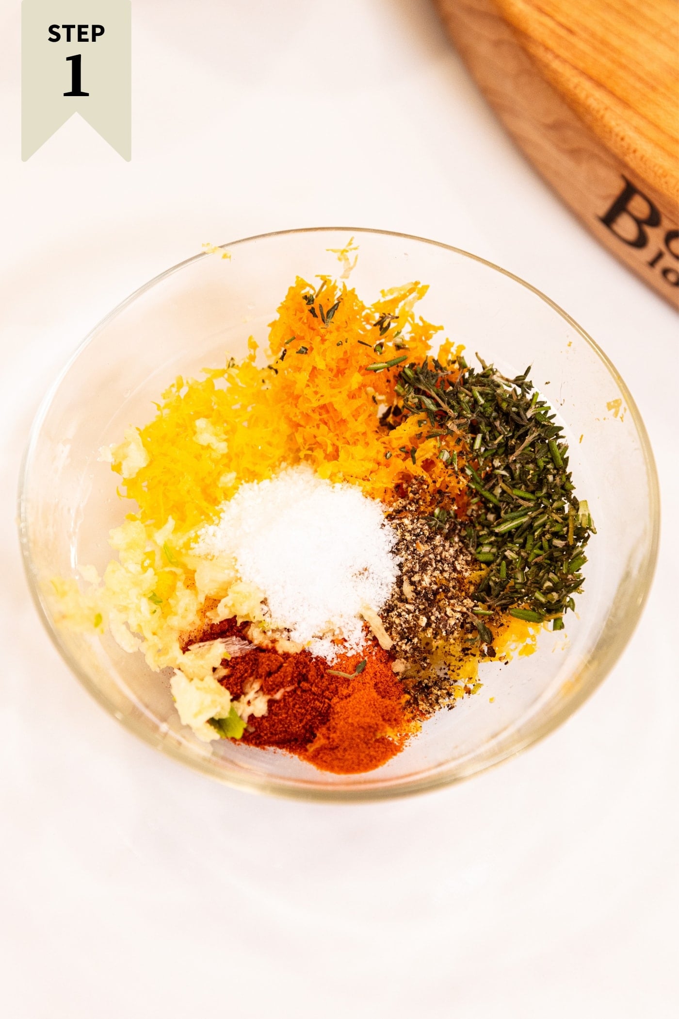 Small glass bowl sitting on white countertop filled with mixture of spices, herbs, and salt before being mixed together with wood cutting board in background.