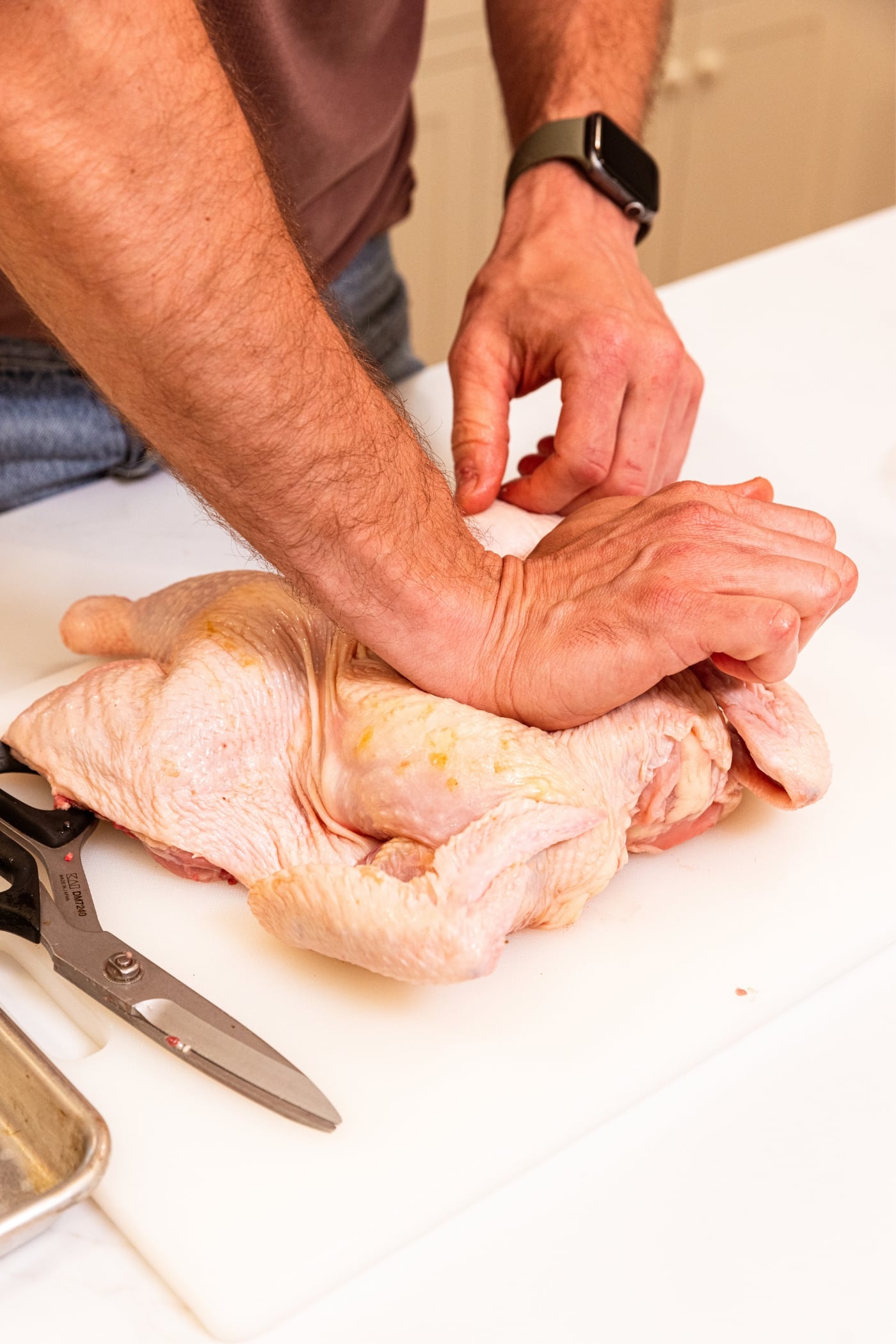 Hand using palm to smash a raw chicken to spatchcock it on a white cutting board.