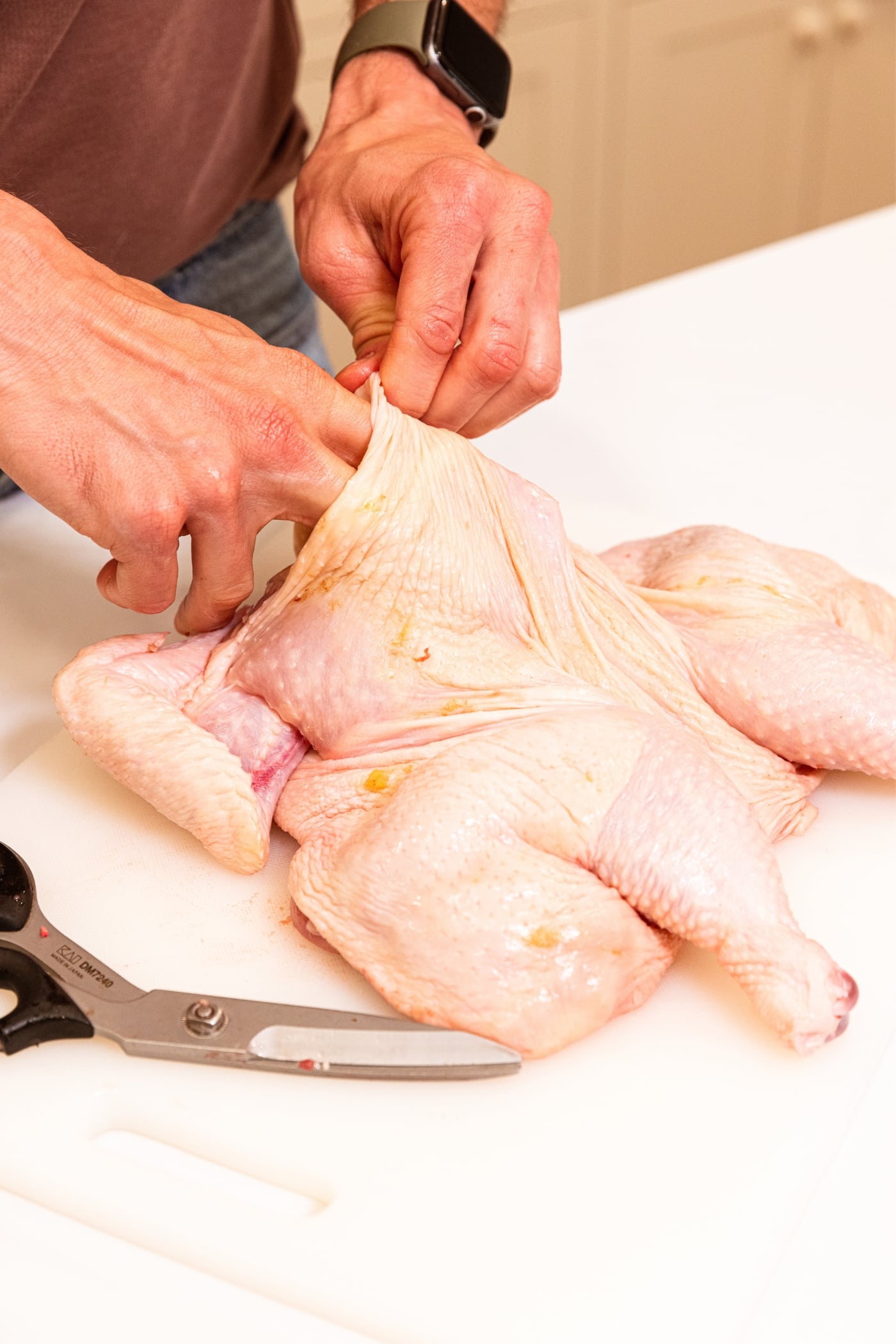 Two hands using fingers to stretch the skin from the body of a raw chicken sitting on white cutting board.