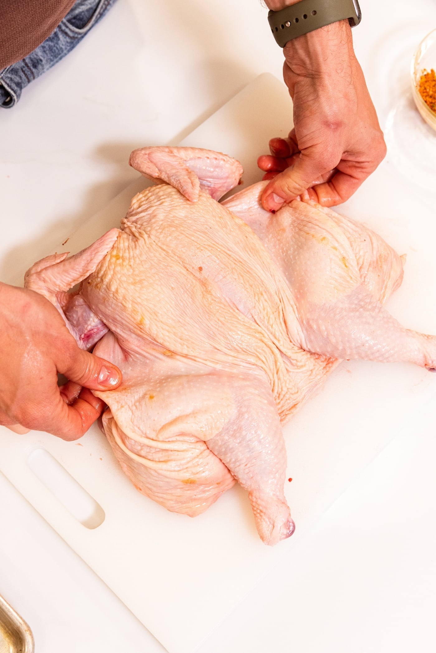 Two hands placing raw chicken on white cutting board that has been spatchcocked.