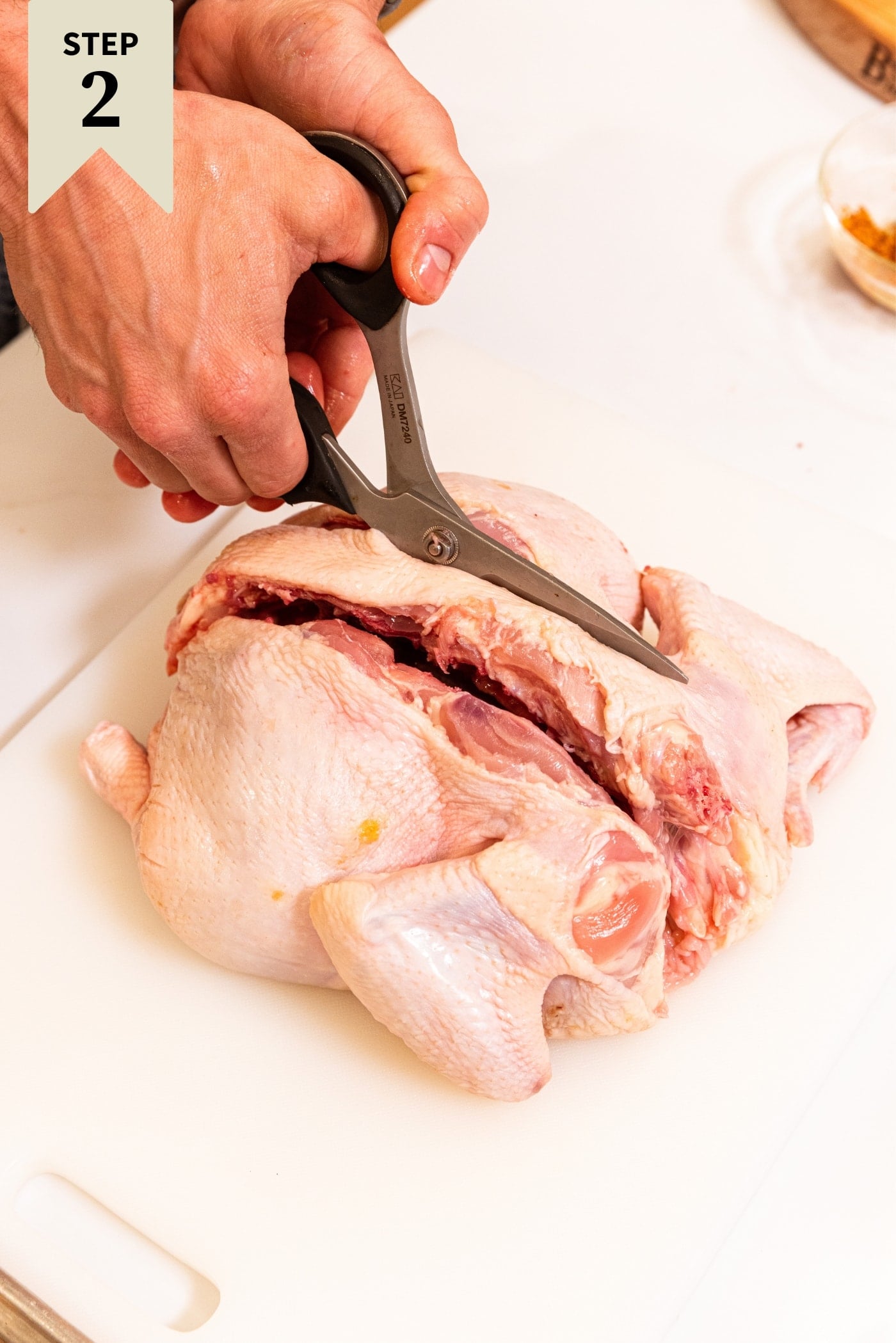 Raw chicken sitting on white cutting board with hand using scissors to cut out the backbone of the chicken.