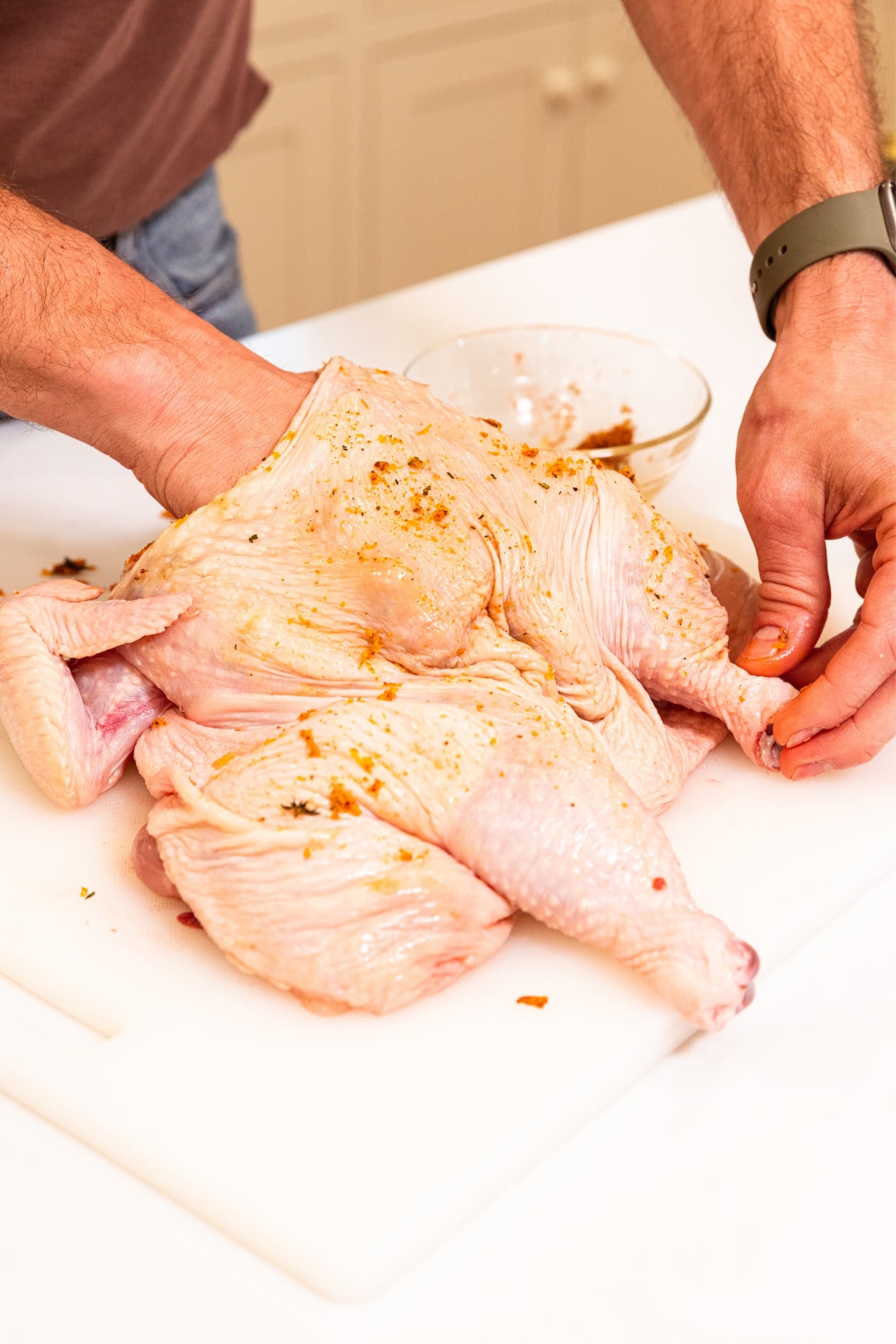 White cutting board with raw chicken sitting on top with spice rub on top placing the spice rub also under the skin.
