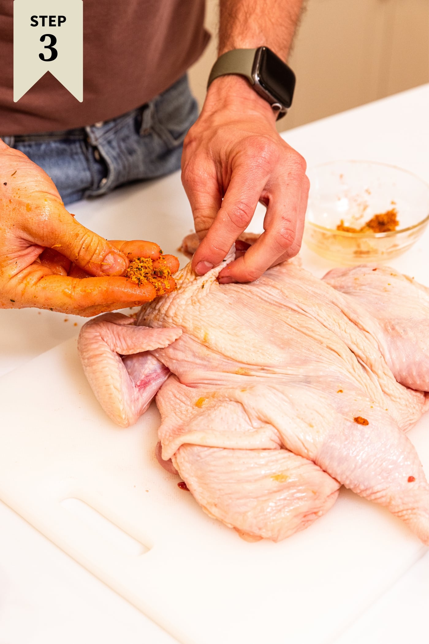 Hand holding spice and salt mixture about to place under the skin of a raw chicken sitting on a white cutting board.