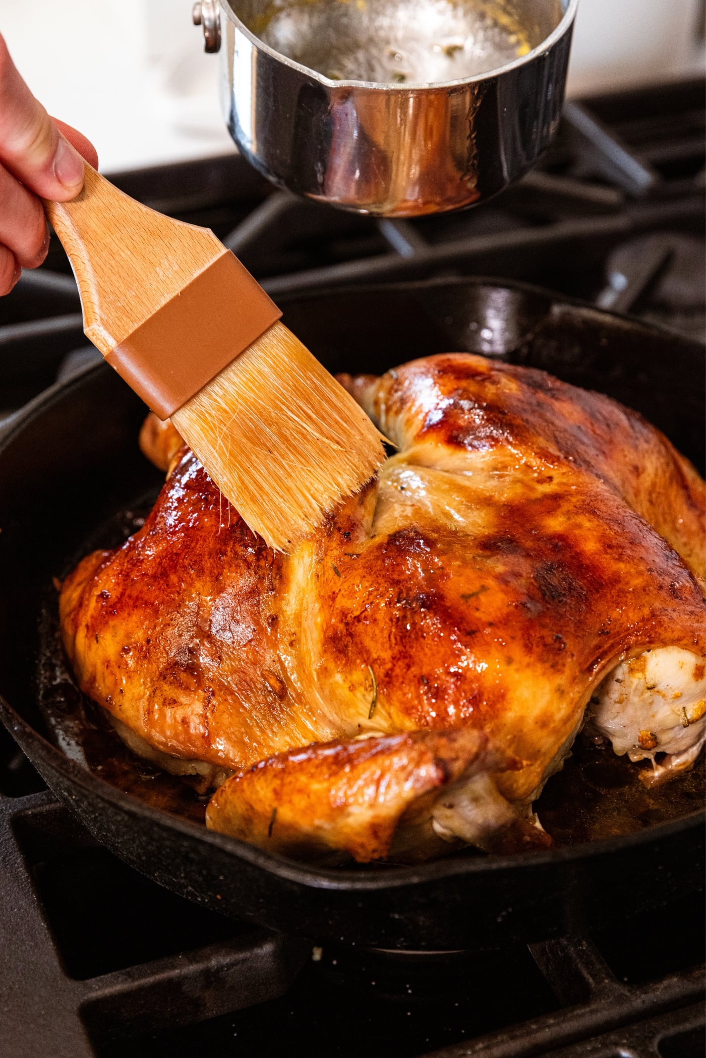 Black skillet with roast chicken sitting in it brushing butter over the top to make it even more golden.