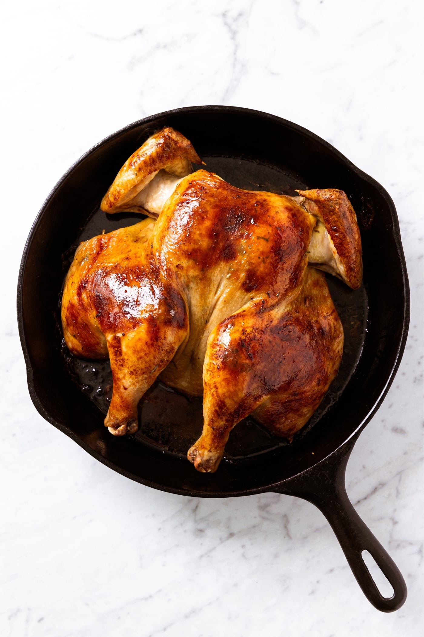 Black skillet sitting on white marble surface with roast chicken right after coming out of the oven with golden top.