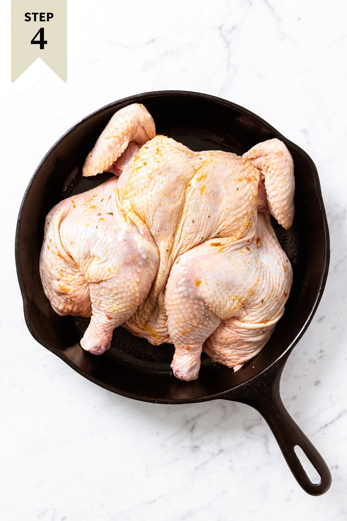 Black skillet sitting on marble surface with raw spatchcocked chicken with spice rub on top before going into the oven to roast.