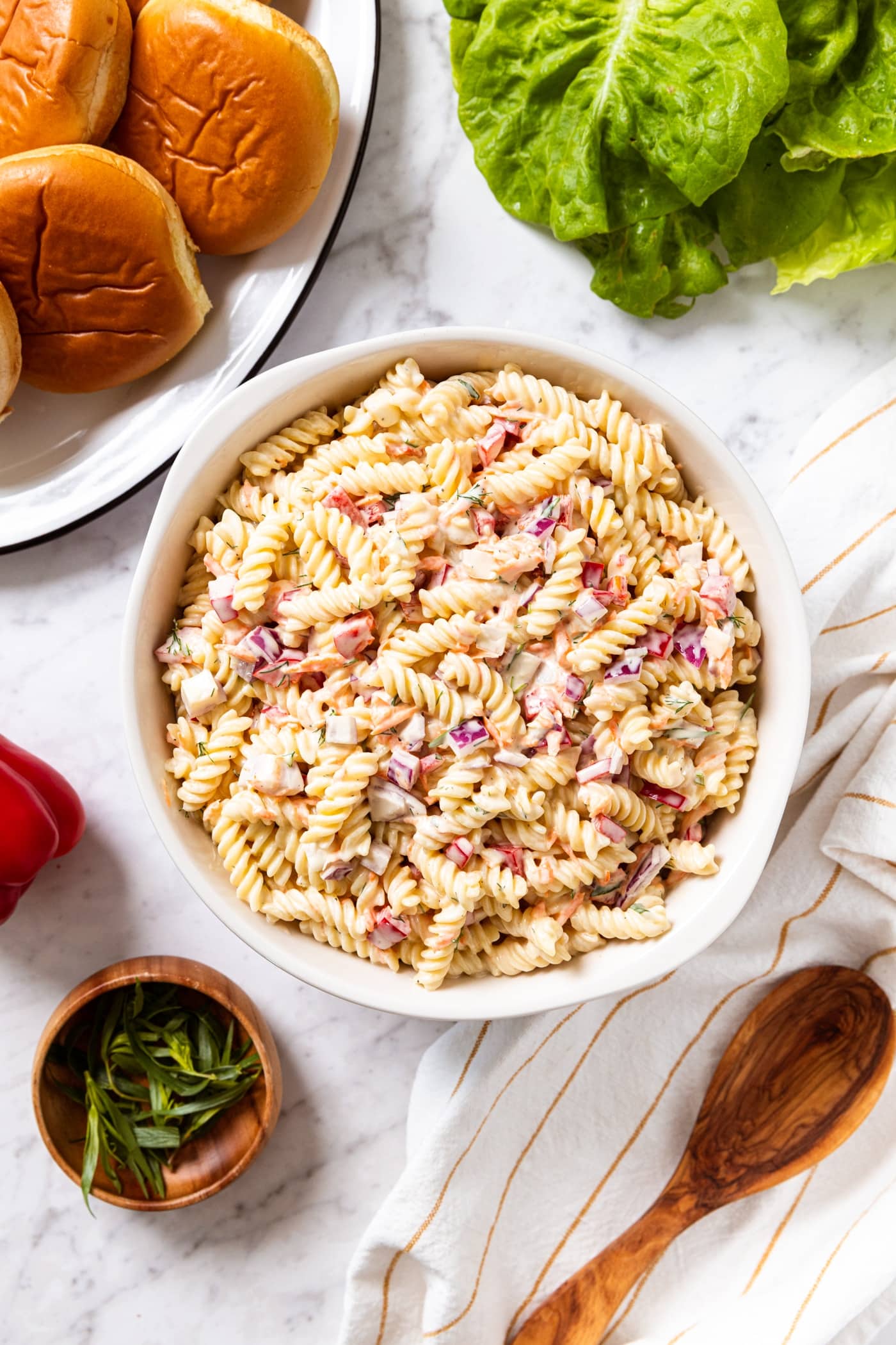 Marble surface with large bowl filled with creamy pasta salad with lettuce, hamburger buns, red bell pepper, herbs, and a wooden spoon.