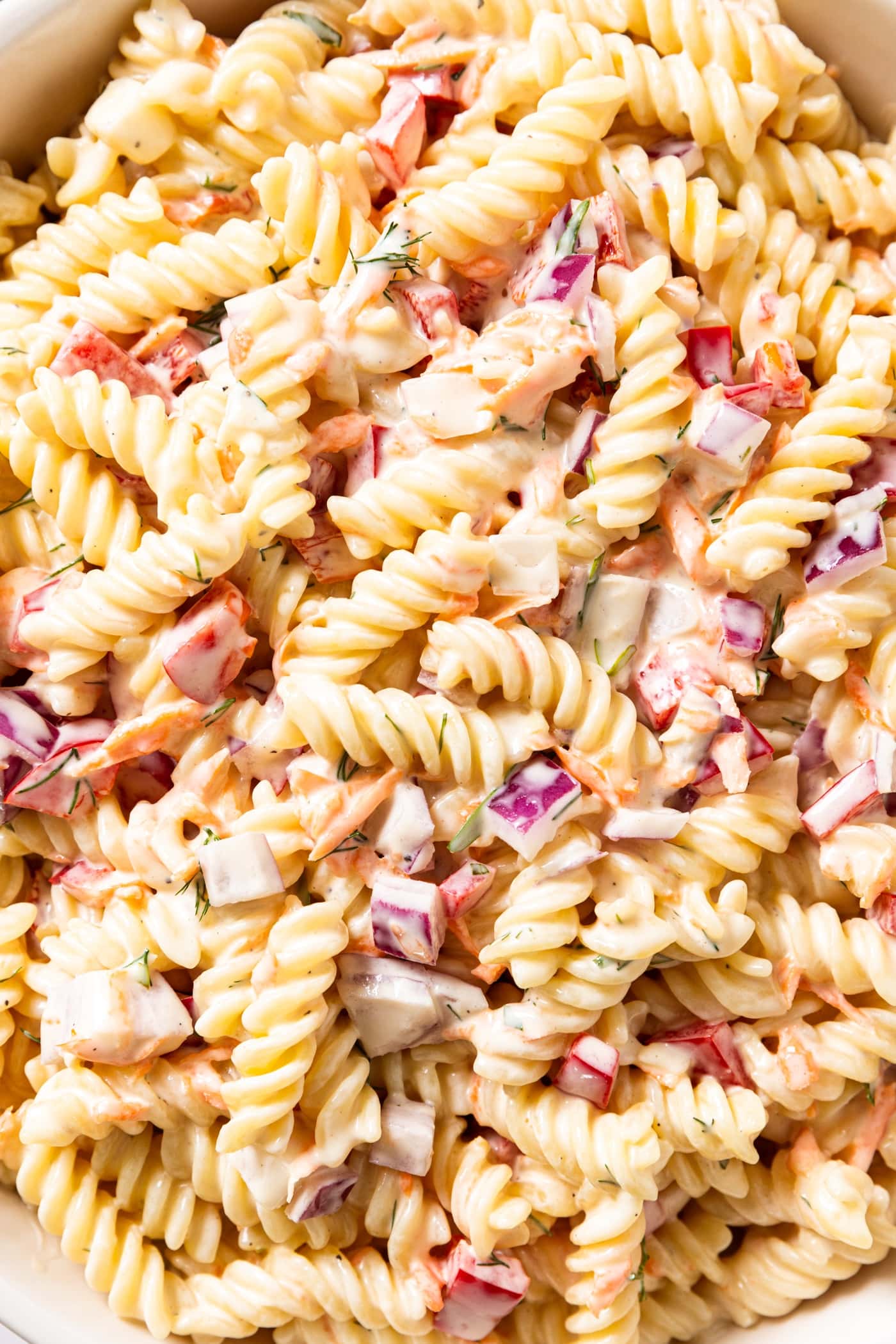 Cooked pieces of pasta covered in creamy dressing tossed with pieces of red pepper, red bell pepper, and grated carrot.