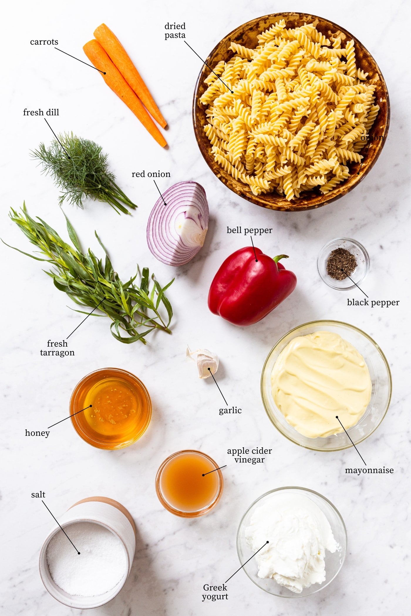 Marble surface with all ingredients needed to make creamy pasta salad including dried pasta, carrots, herbs, red onion, bell pepper, honey, mayonnaise, and more.
