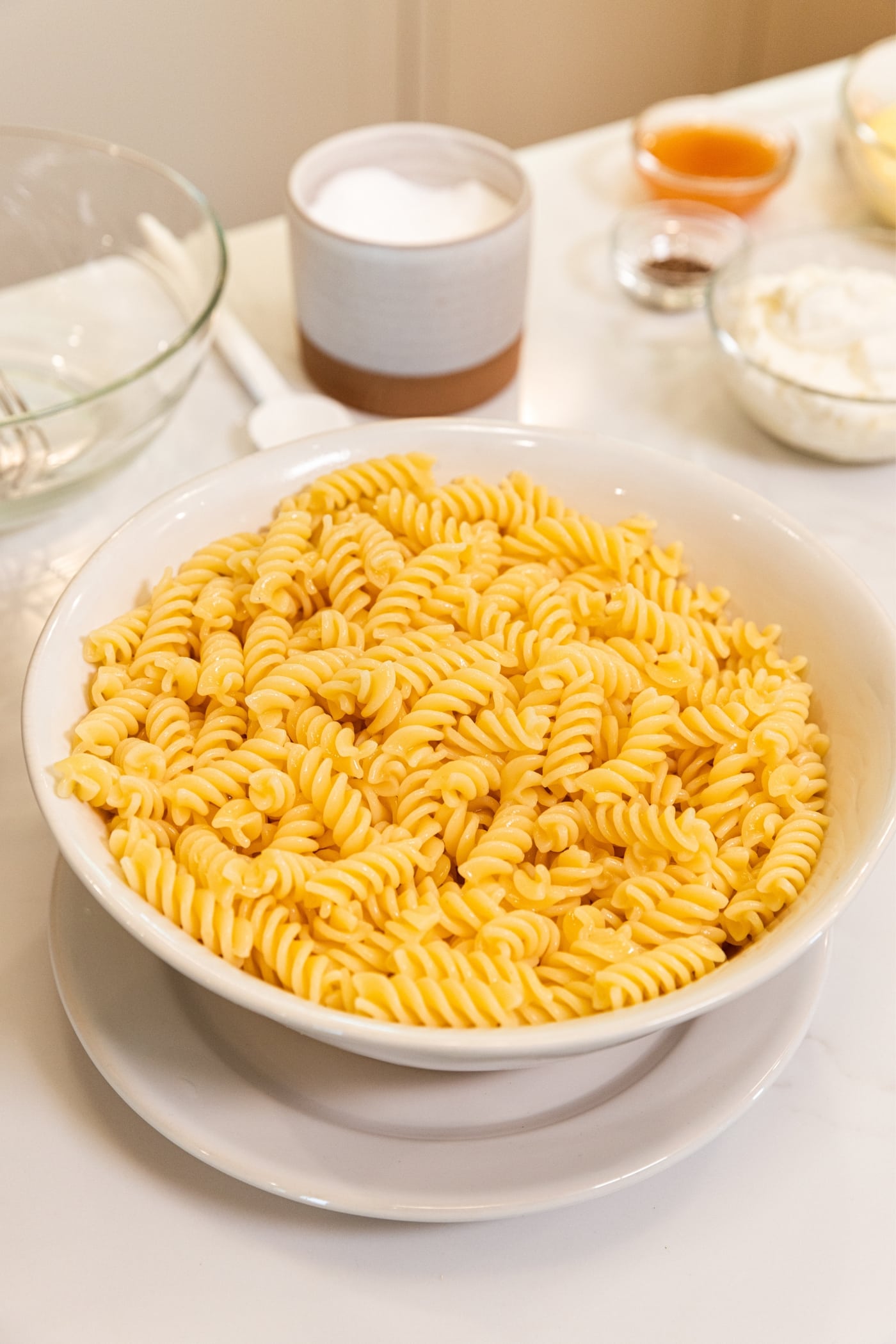 White colander with cooked pasta draining in bowl on white plate all on white surface with other ingredients in background.
