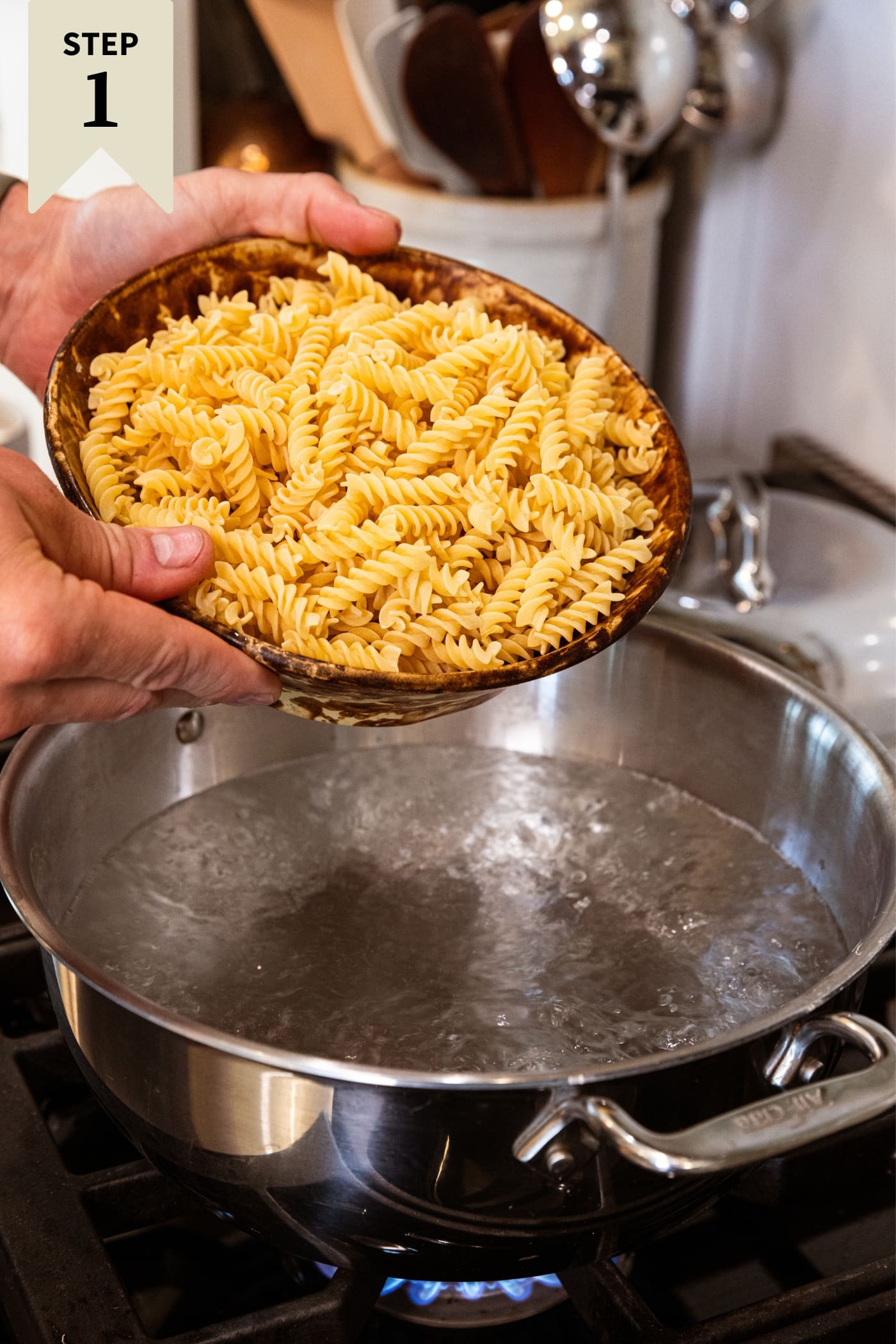 Two hands holding brown bowl about to pour uncooked pasta into a large pot of boiling water on stovetop.