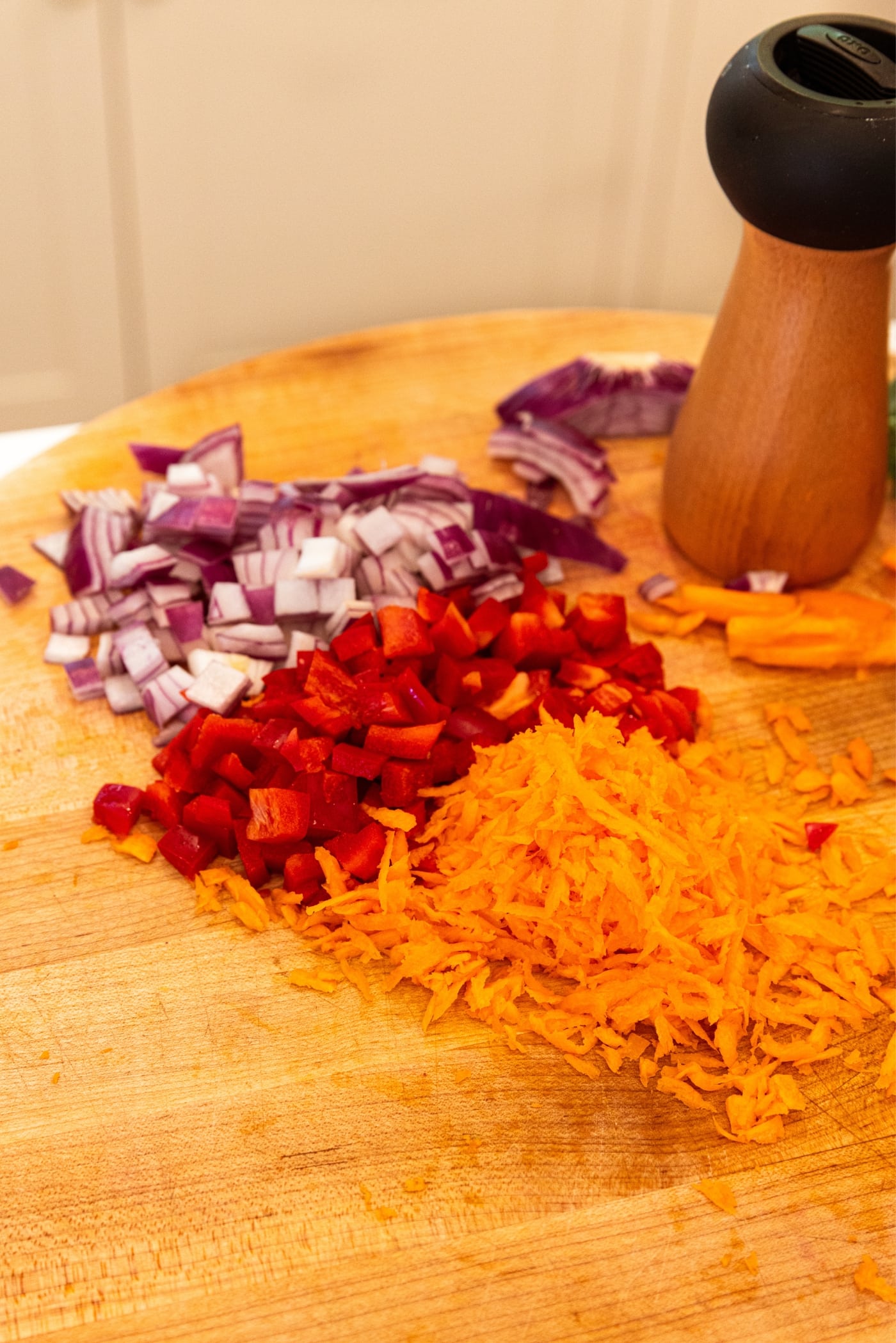 Wooden cutting board with diced red onion, diced red bell pepper, and grated carrot with black pepper grinder in background.