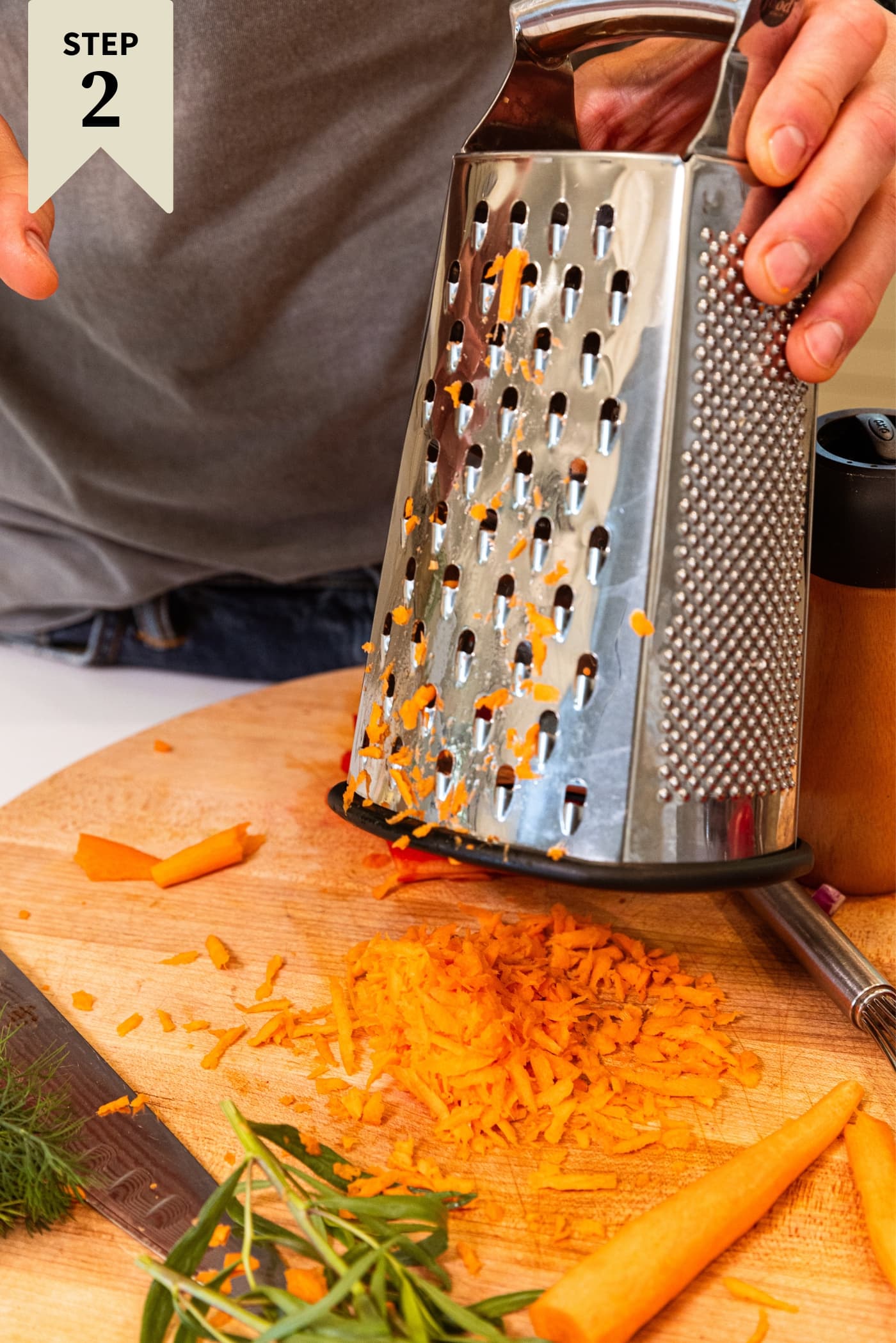 Hand holding box grater holding above grated carrot on wood cutting board.