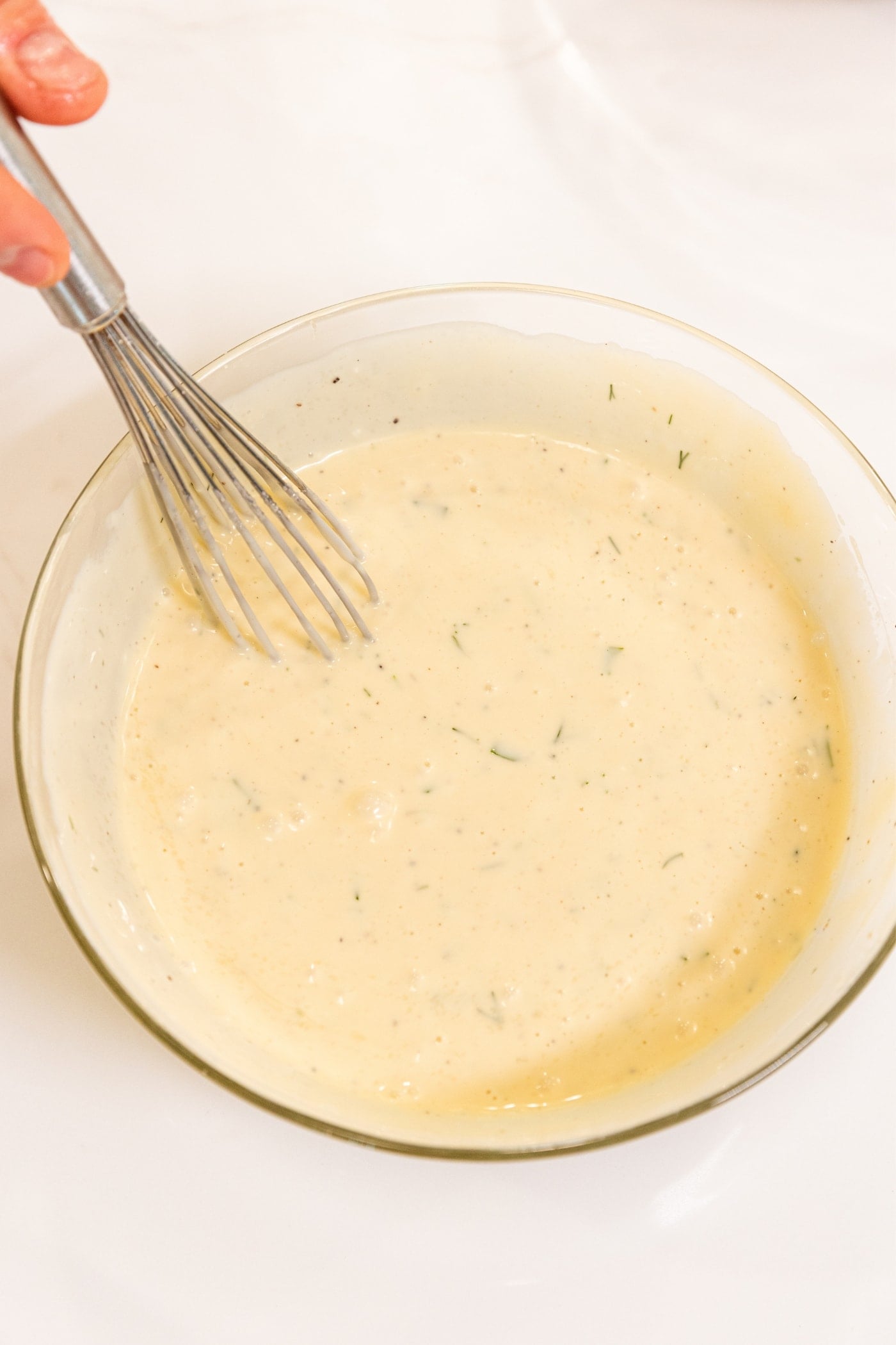 Hand holding wire whisk in middle of small glass bowl filled with cream-colored creamy dressing for pasta salad all on white surface.
