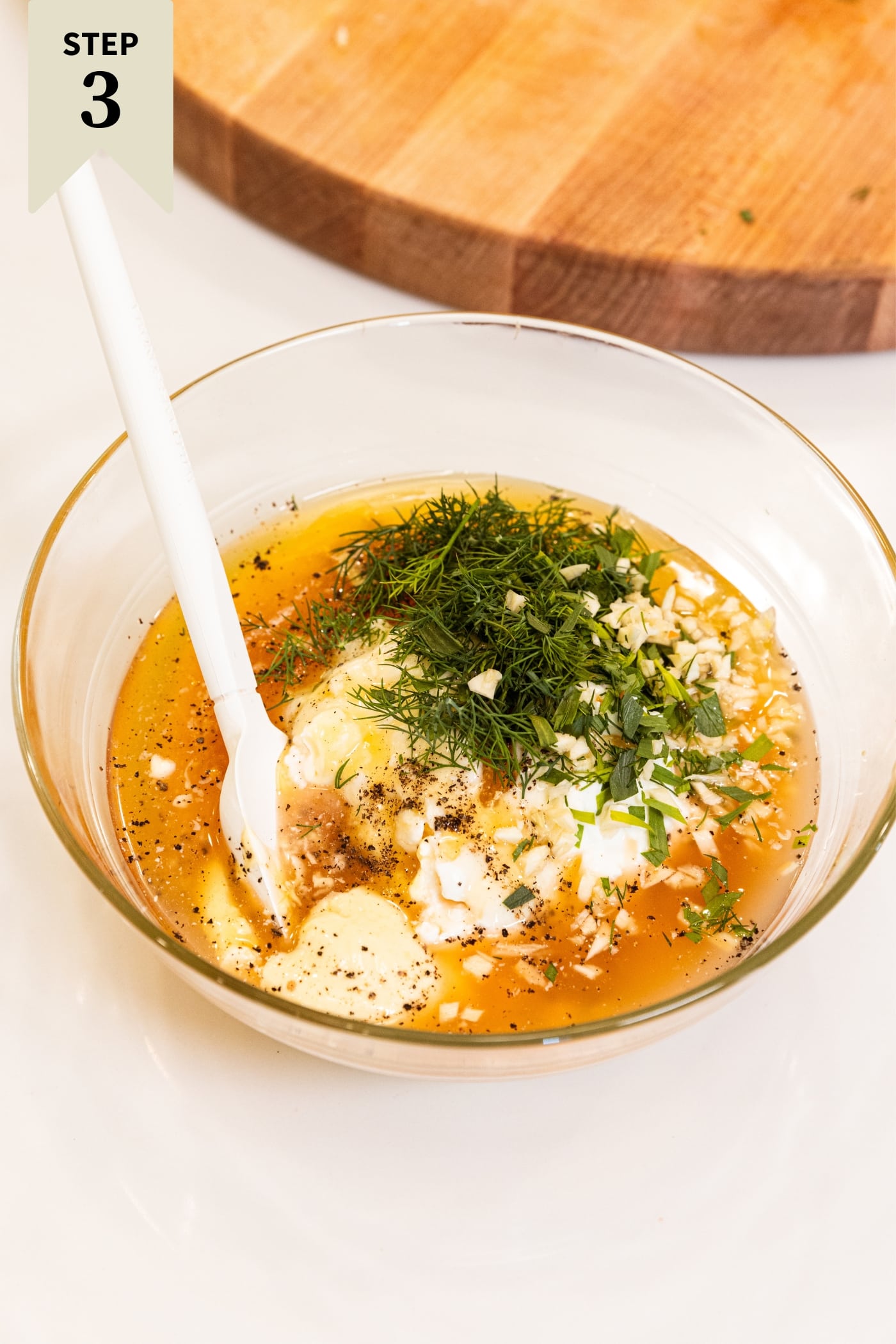 Small glass bowl filled with all ingredients needed to make a creamy pasta dressing including mayonnaise, yogurt, fresh herbs, honey, and apple cider vinegar all on white surface with wood cutting board in background.
